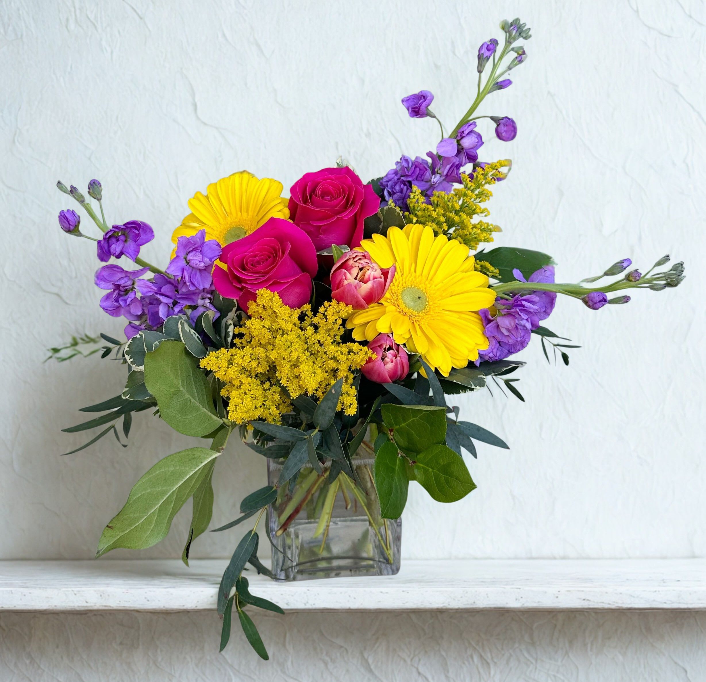 Color Me Happy - A bright and joyful arrangement bursting with vibrant color. Sunny yellow gerbera daisies and golden accents are paired with rich pink roses, soft tulips, and delicate purple blooms for a fresh, garden-inspired look. Designed in a clear glass cube with lush greenery, this cheerful bouquet brings warmth and happiness to any space—perfect for celebrations or simply brightening someone’s day.