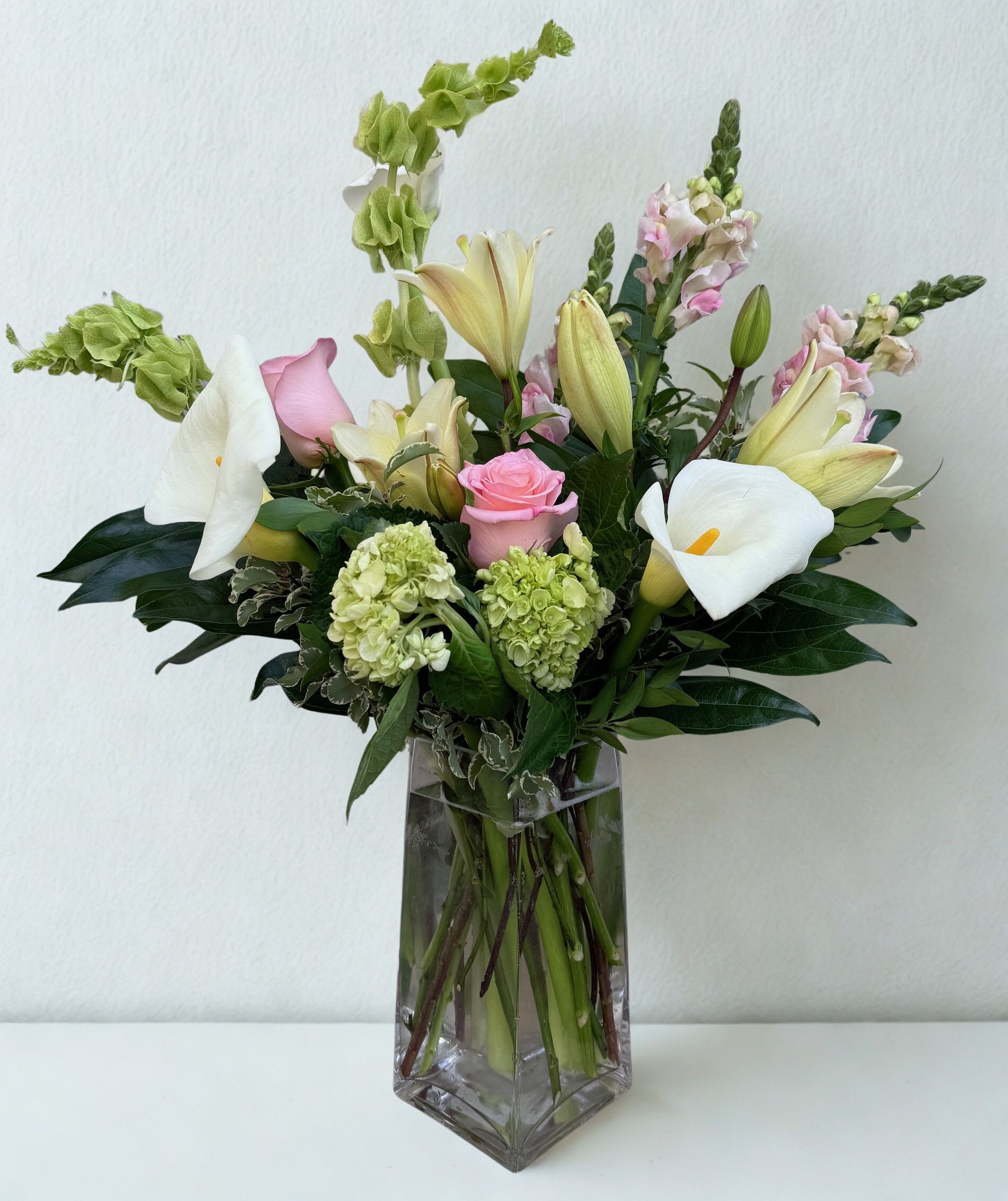 Pastel Promise - refined and elegant arrangement featuring graceful white calla lilies, soft blush roses, and creamy lilies accented with delicate snapdragons and lush green hydrangea. Designed in a sleek glass vase with rich greenery, this bouquet offers a perfect balance of softness and sophistication. Ideal for expressing heartfelt sentiments, celebrating special moments, or adding a touch of timeless beauty to any space.