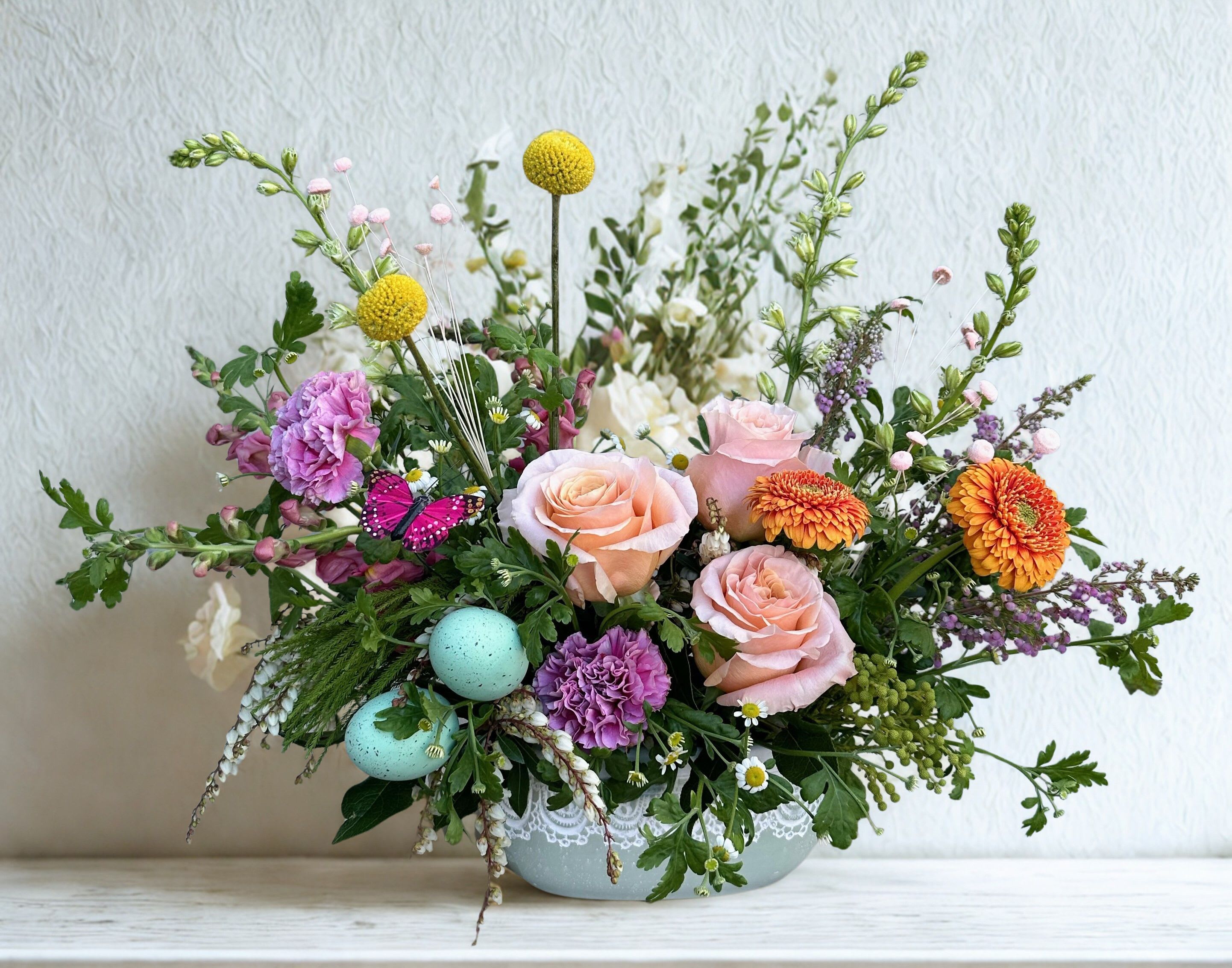 Robin's Garden Delight - A cheerful spring arrangement bursting with soft peach roses, vibrant ranunculus, and playful pops of lavender and yellow blooms. Nestled among lush greenery, delicate speckled eggs and a whimsical butterfly add a touch season charm. 