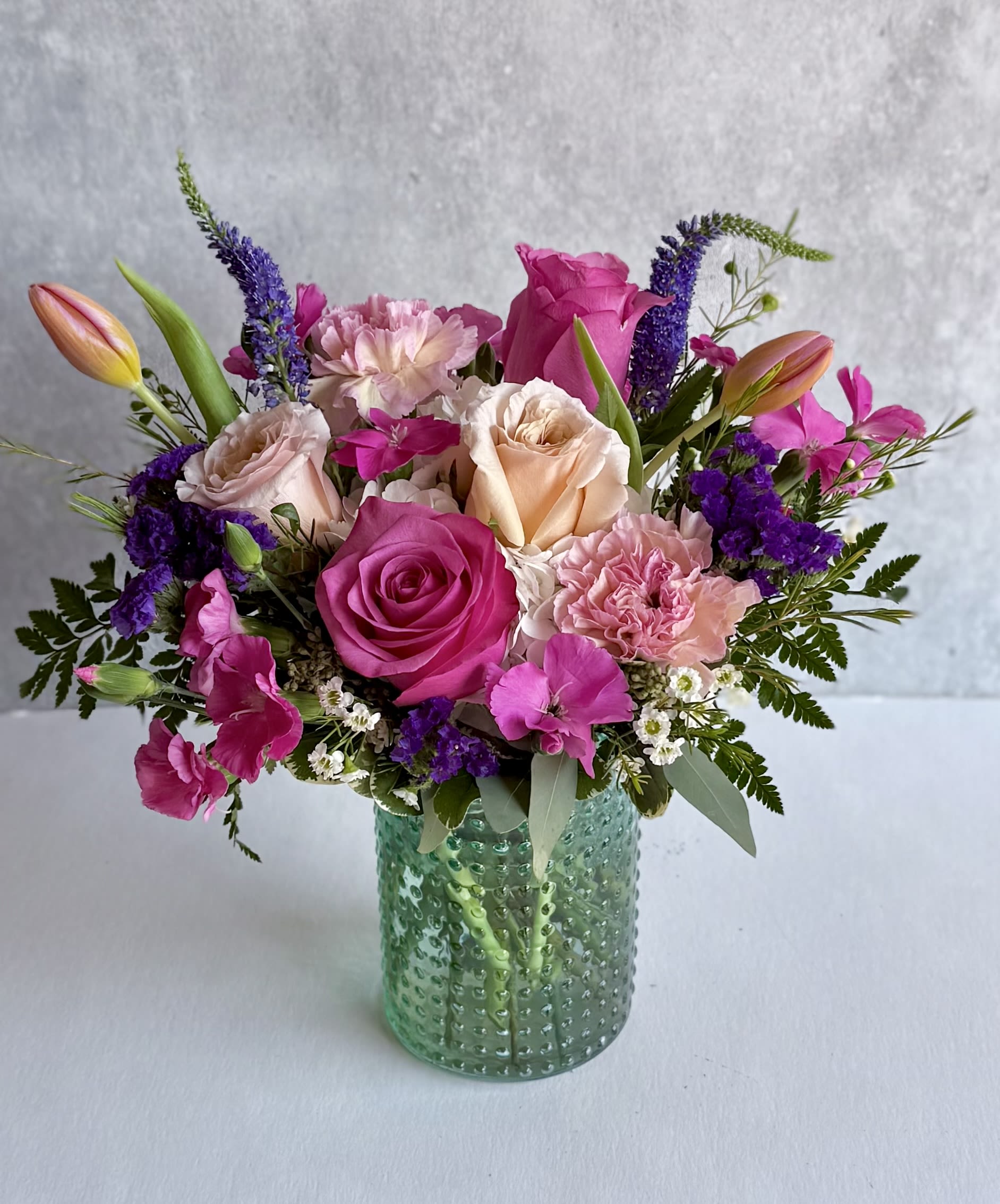 Mixed bouquet of pink roses, carnations, and tulips with purple accents in a textured green glass vase