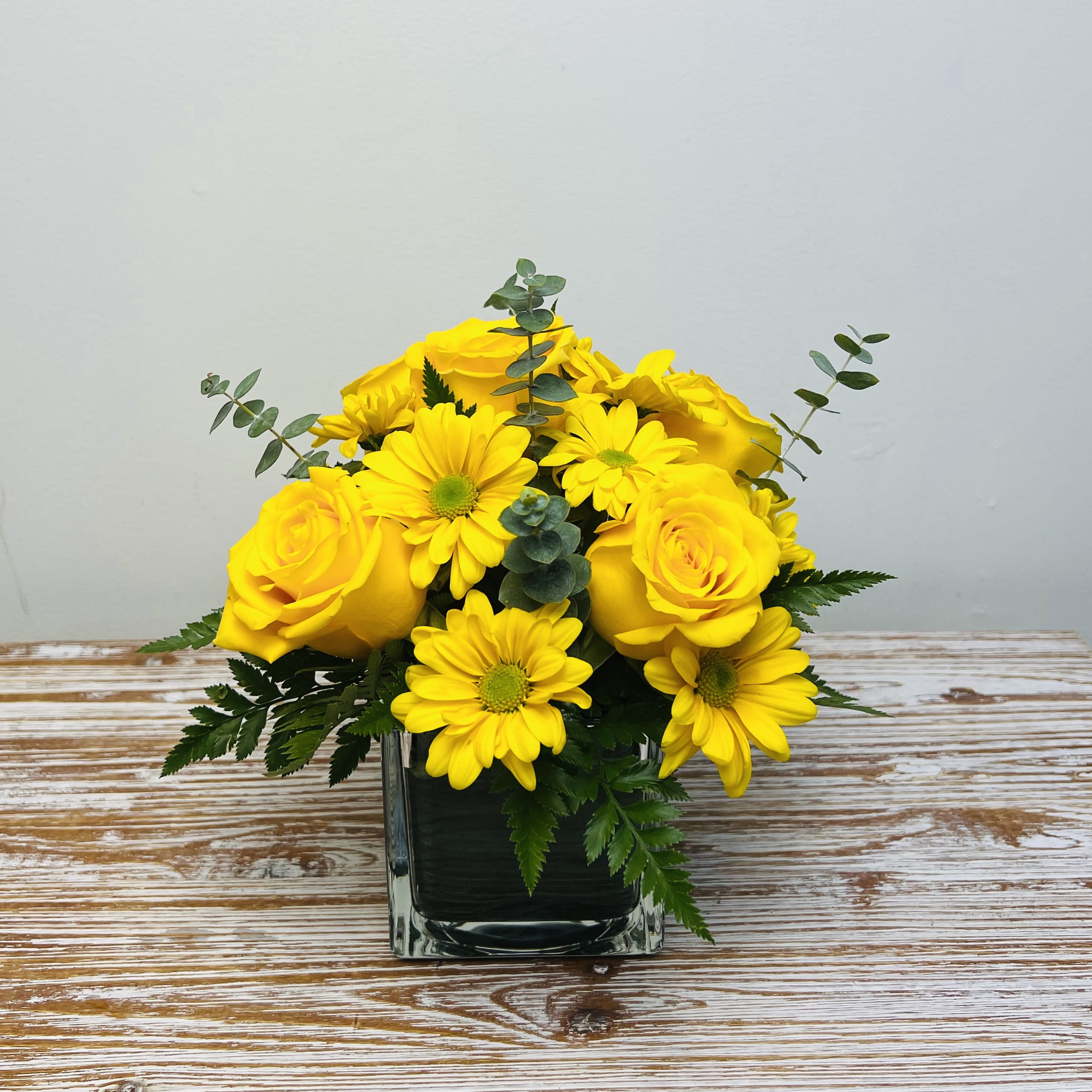 Sunshine - This arrangement is full of bright bold yellows of roses and spray daisies, accented with eucalyptus in a leaf wrapped clear cube vase. A great choice to cheer someone up!  Approximately 10" Tall and 8" Wide  Container Country of Origin: Mexico Flower Country of Origin: Ecuador and/or Columbia