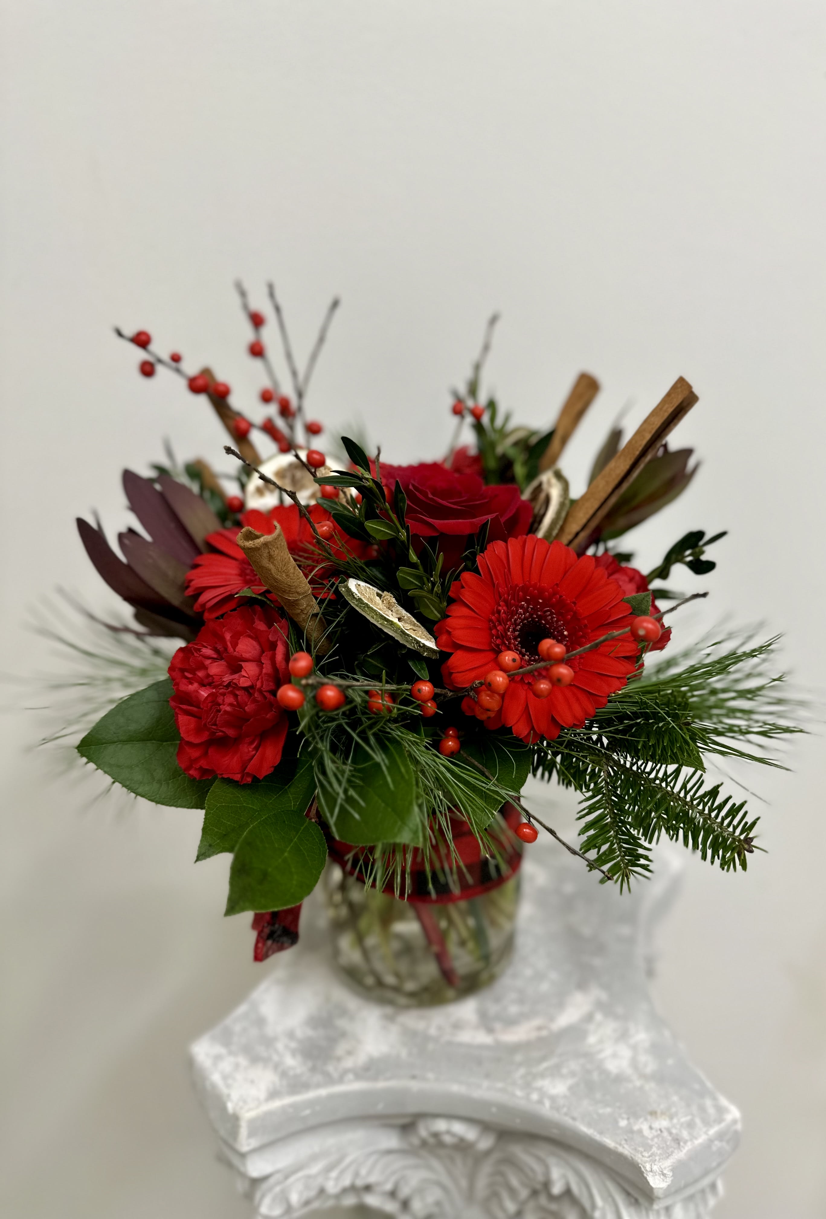 Cinnamon Spice by Westford Florist Art by Veny - Indulge in the coziness of the season with Cinnamon Spice by Westford Florist Art by Veny. This stunning display showcases a perfect combination of deep red roses, lively gerbera daisies, and graceful carnations, accompanied by the charming allure of safari sunset blooms and plump red winter berries. Each arrangement is thoughtfully accented with fragrant cinnamon sticks and tangy dried lemon slices, capturing the magic of the holidays in every aspect. Add a touch of sophistication to your home or delight a loved one with this captivating centerpiece that effortlessly blends elegance and seasonal cheer. Experience every moment as special with the inviting warmth of Cinnamon Spice.