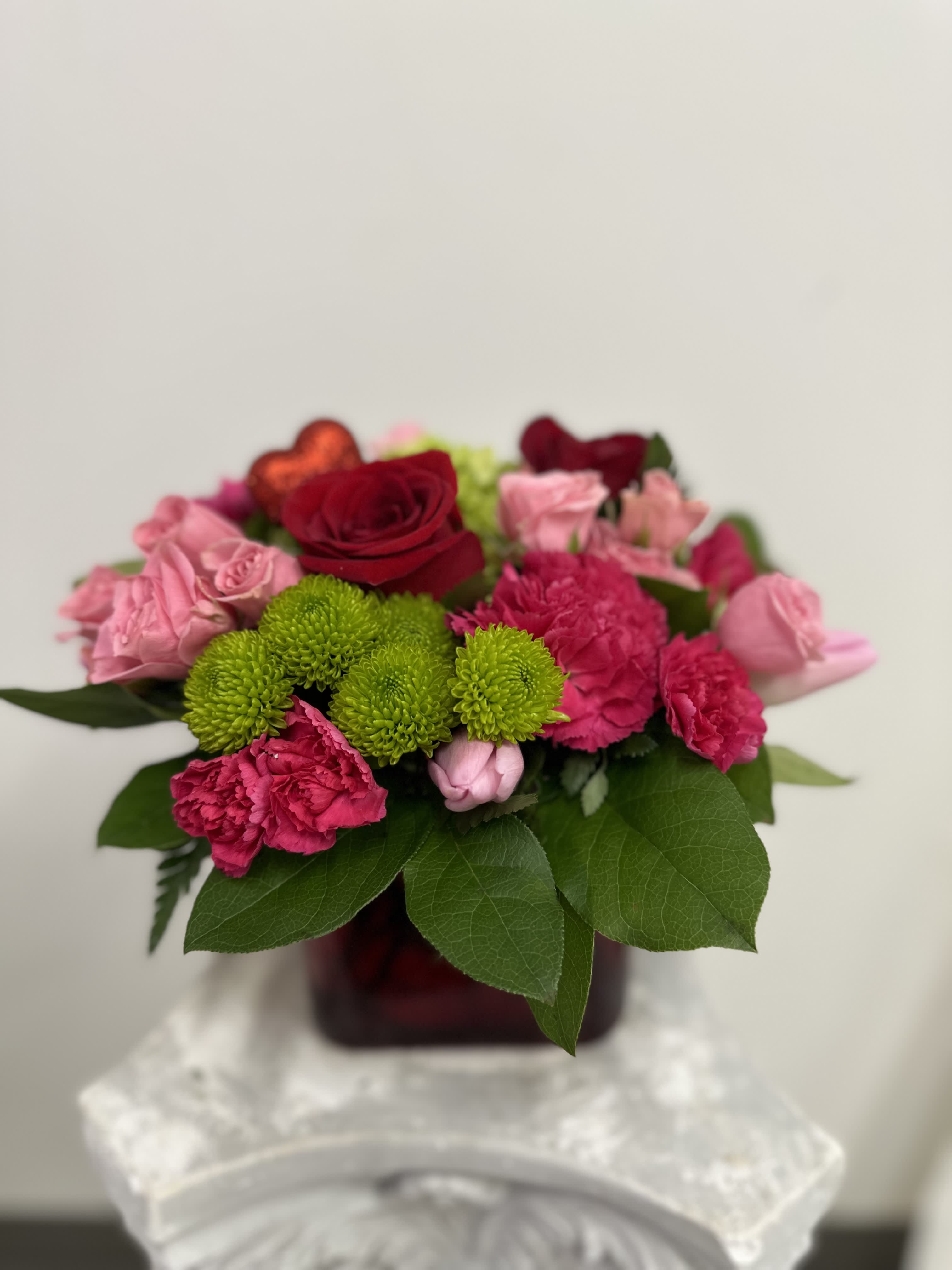 Amour by Westford Florist Art by Veny - Elevate your gifting experience with Amour by Westford Florist Art by Veny. This exquisite arrangement features a harmonious blend of lush red and soft pink blooms, artfully complemented by vibrant greens, all perfectly housed in an elegant red petite cube vase. Ideal for expressing love or celebrating special moments, this stunning floral display radiates romance and sophistication. Each piece is meticulously crafted to bring warmth and beauty into any space, making it the perfect choice for anniversaries, heartfelt apologies, or simply to show someone you care. Experience the art of floral elegance with Amour—a timeless bouquet that speaks volumes without saying a word.