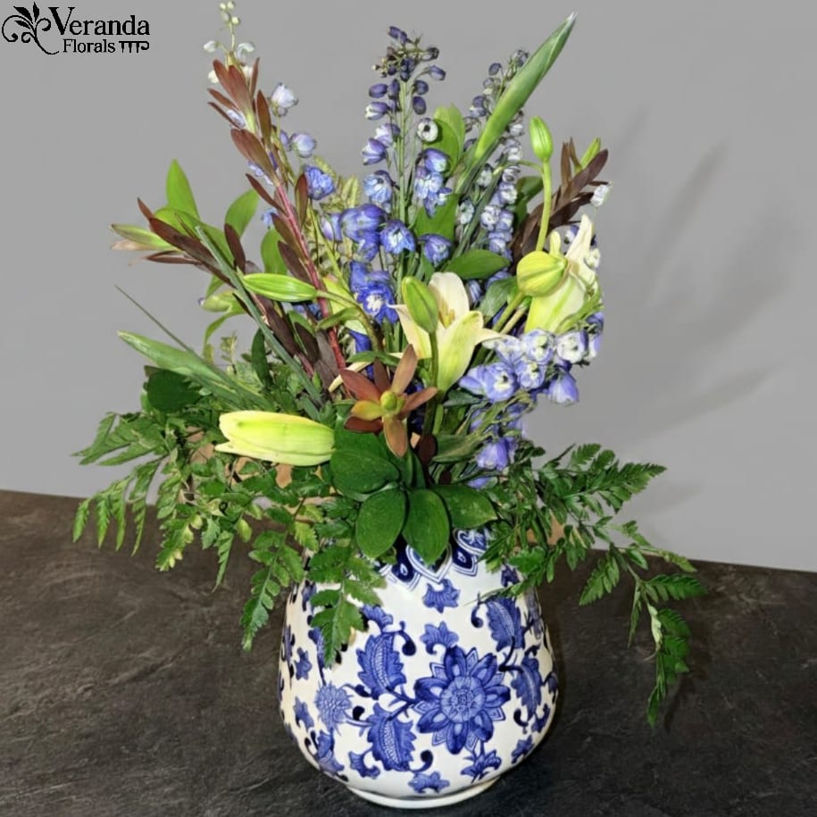 Blue And White Wide Bottom Vase - Elevate your home décor with this stunning blue and white Chinoiserie-style vase. A timeless classic, this piece features intricate cobalt blue floral patterns hand-painted onto a crisp white porcelain-style base. The delicate vine motifs and bold blooms offer a sophisticated aesthetic that seamlessly blends traditional charm with modern elegance.  Price includes the vase and designers choice of flowers. 