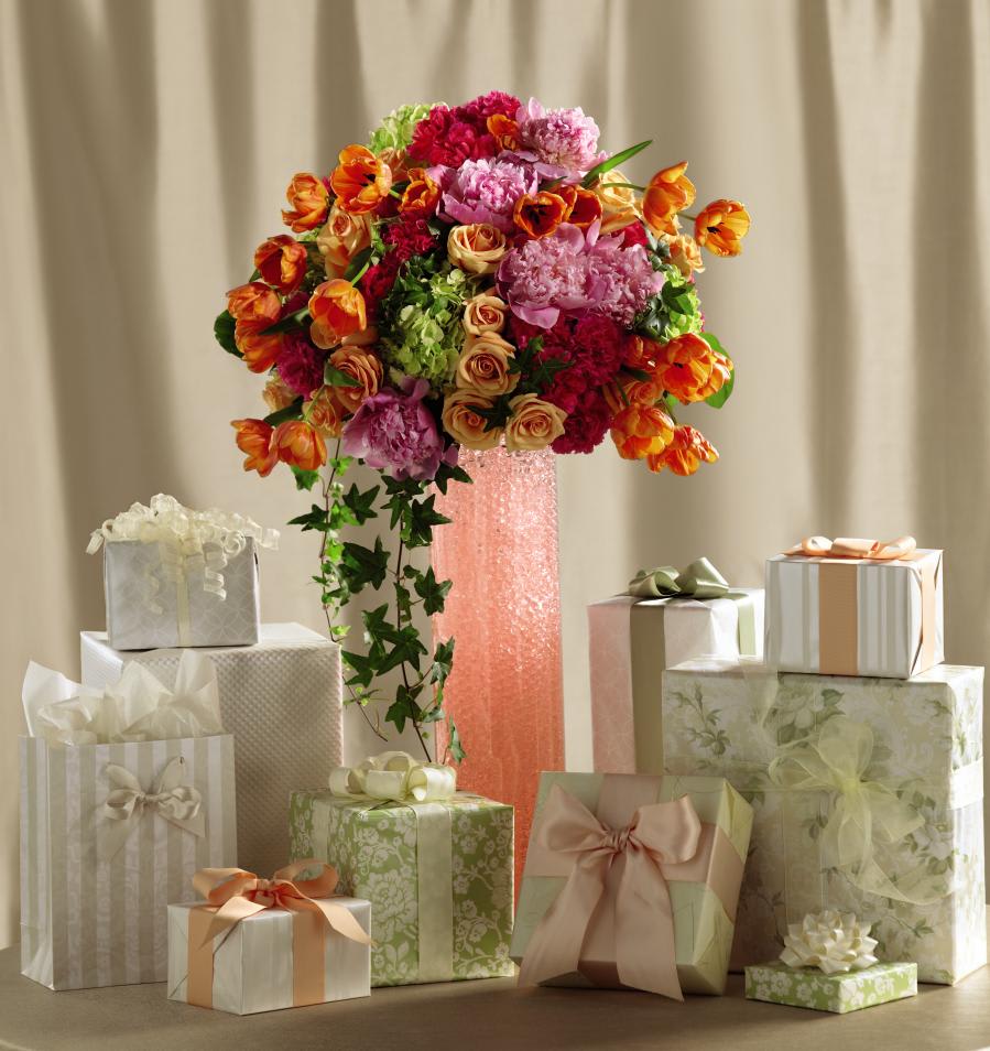 Tall pink vase arrangement of orange tulips, pink peonies, and roses surrounded by pastel gift boxes