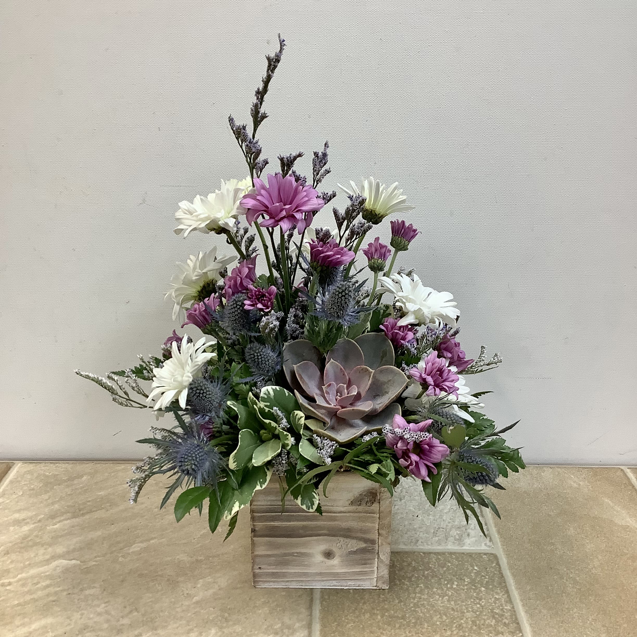 Lavender - This floral arrangement has a rustic, natural charm with a soothing lavender-inspired palette. It's displayed in a weathered wooden box, giving it an earthy, farmhouse feel. A rosette-shaped succulent in dusty green and mauve tones is surrounded by white and lavender-hued daisies, blue thistle and blue limon accent flowers. Variegated greenery adds lushness and depth.   Approximately size: 12” wide x 12” tall.  Primary flowers: daisy, succulent, thistle