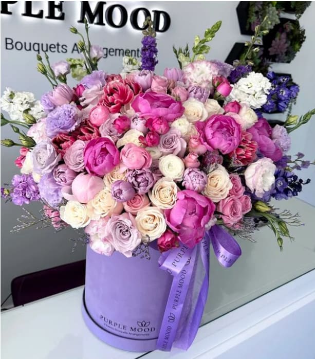 Large round arrangement of pink, lavender, and white blooms in a lavender hat box with ribbon.