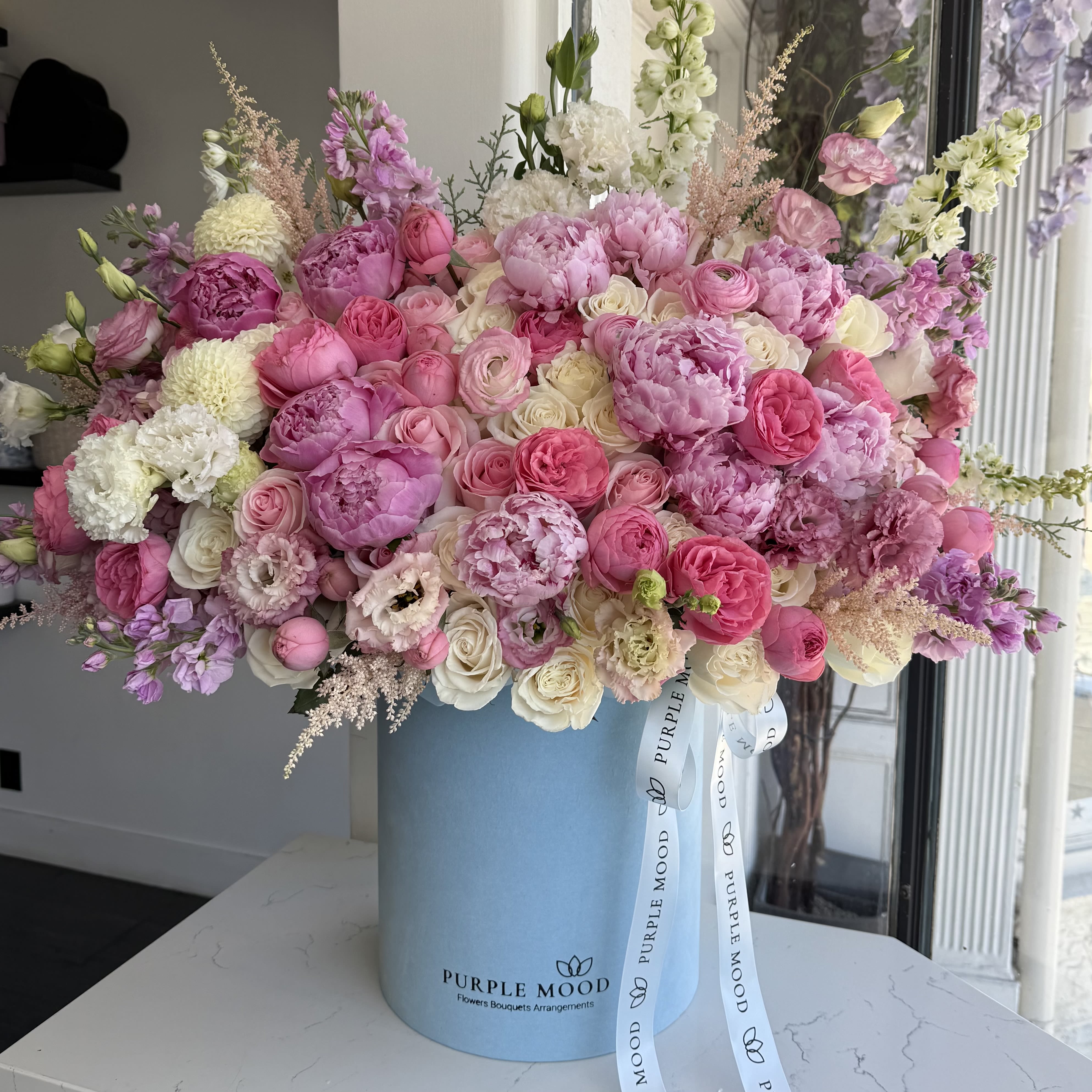 Blue Dream Box - Container size D10.5'x H12.25'  Overflowing with delicate peonies, spray roses, lisianthus, astilbe, and garden roses in soft pinks, creams, and lavenders, this lush arrangement is the definition of romantic elegance. Designed in our signature sky-blue hat box, the Blue Dream Box is perfect for celebrating love, beauty, and unforgettable moments. 