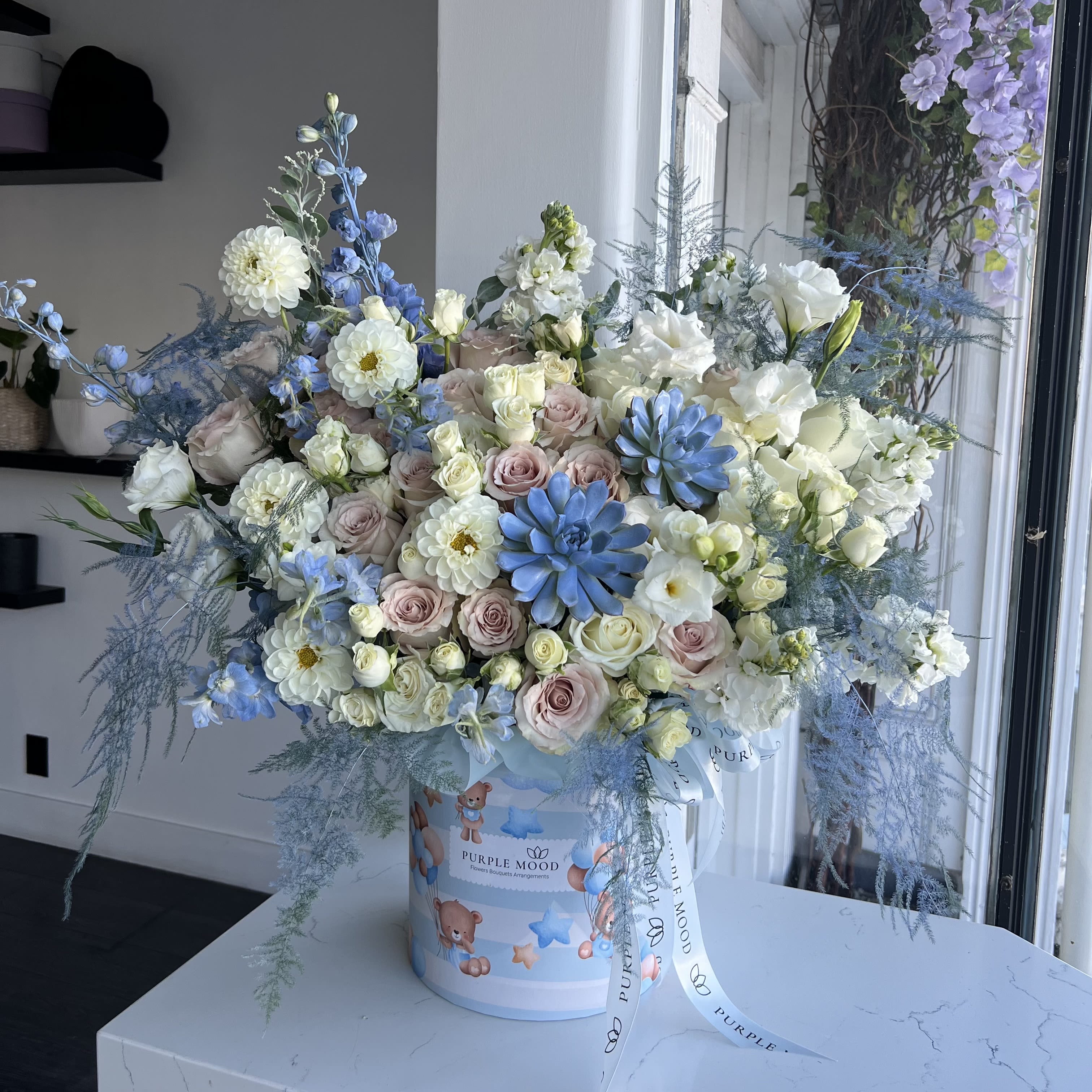 Boy Box - Deluxe size same as pictured. Container size D8.25’x H9.5′. Front side arrangement with lid in the back.  Celebrate the arrival of a precious baby boy with our charming Boy Box arrangement. This elegant design features a soft and soothing mix of pastel roses, white daisies, delicate lisianthus, and fresh greenery, highlighted by unique blue accent blooms. Arranged in a keepsake baby-themed box, it’s the perfect gift for welcoming a little one into the world with love and joy.   •    Premium roses, daisies, lisianthus, and seasonal accent flowers     •    Blue floral highlights for a soft baby-boy theme     •    Presented in a baby-themed decorative box with ribbon     •    Symbolizes purity, new beginnings, and celebration     •    Perfect for baby showers, hospital deliveries, or welcoming home