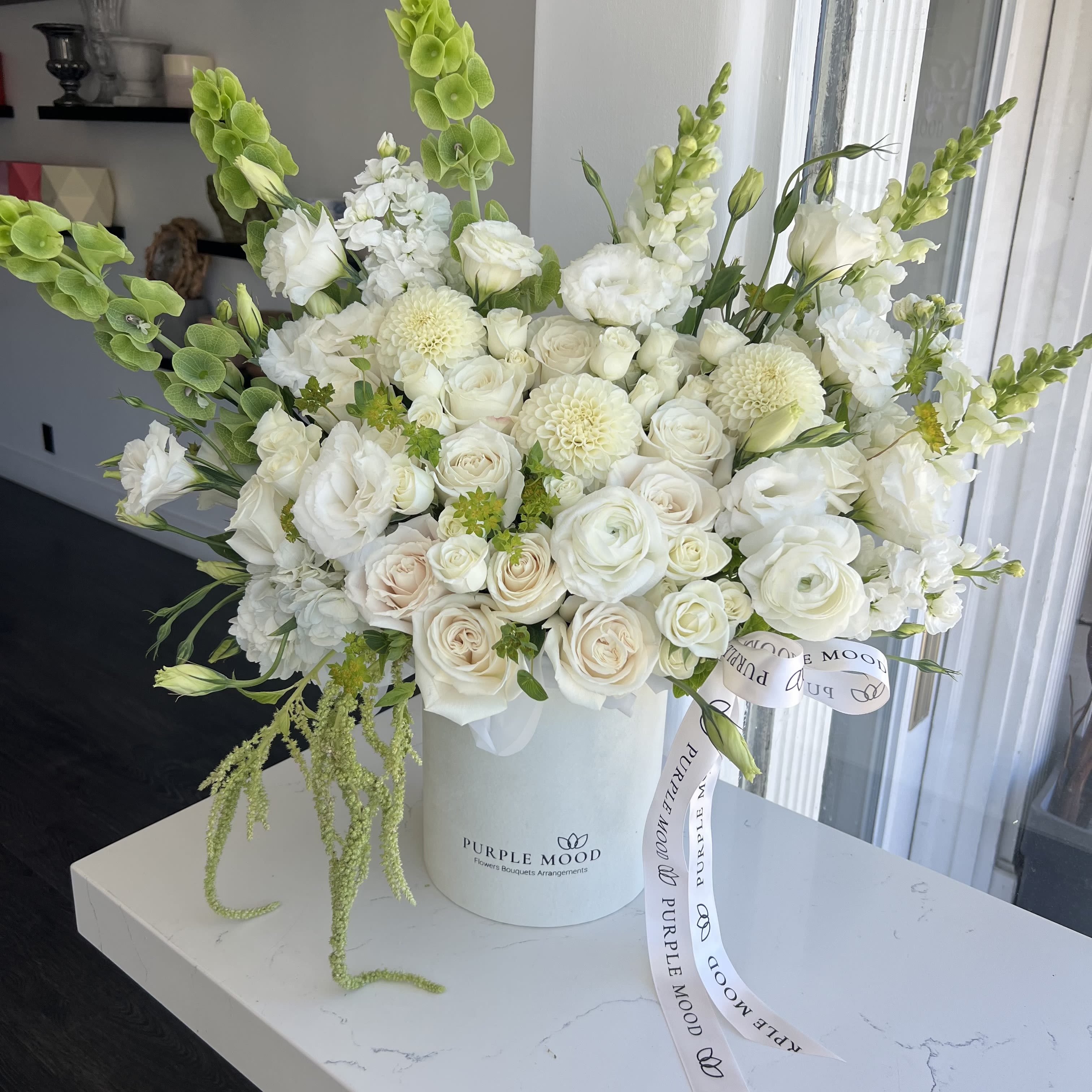 Elegance Box - Container size D6.75'x H7.5'  The Elegance Box is a timeless arrangement that exudes sophistication and charm. Designed in a chic white hat box, this bouquet features a graceful mix of white roses, lush chrysanthemums, and snapdragons, accented with fresh greenery. Its refined monochromatic palette makes it perfect for weddings, anniversaries, or any occasion where you want to convey pure elegance.   •    Includes premium white roses, chrysanthemums, and snapdragons     •    Accented with fresh seasonal greenery for added texture     •    Arranged in a stylish white hat box with branded ribbon     •    Ideal for weddings, anniversaries, sympathy, or formal celebrations     •    Size: Standard (as pictured)