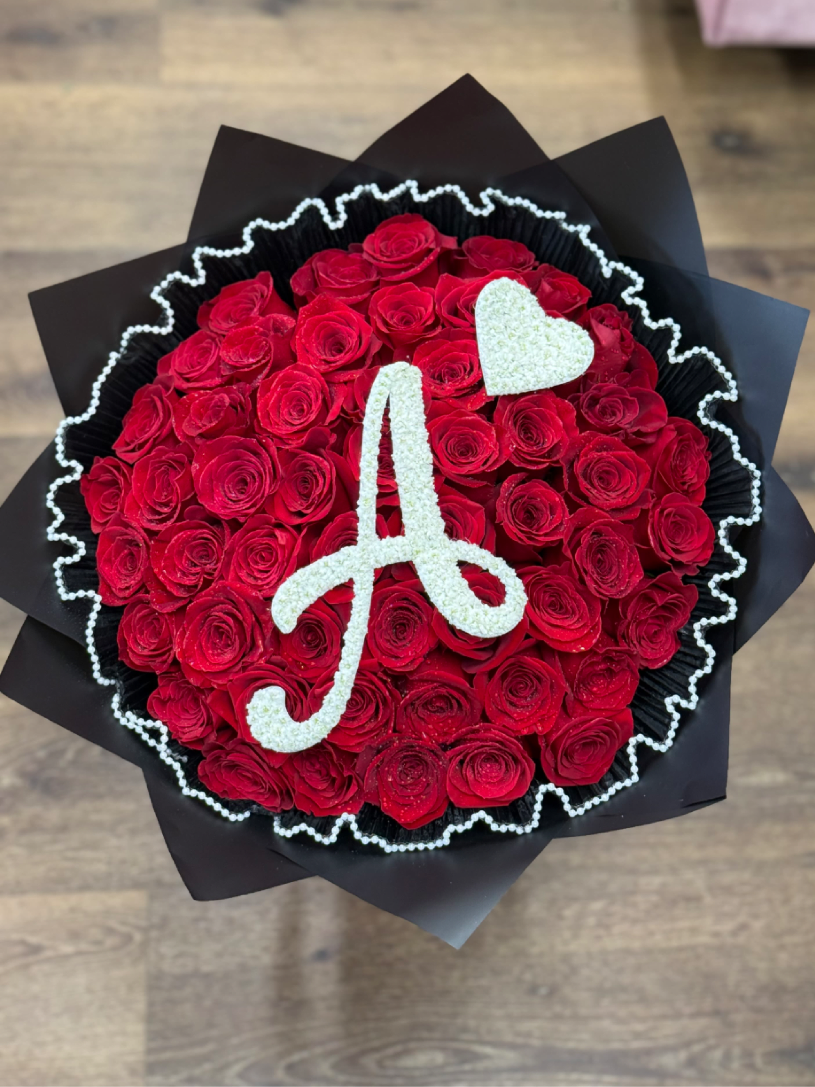 Round arrangement of red roses with a white A initial and heart on top, wrapped in black paper