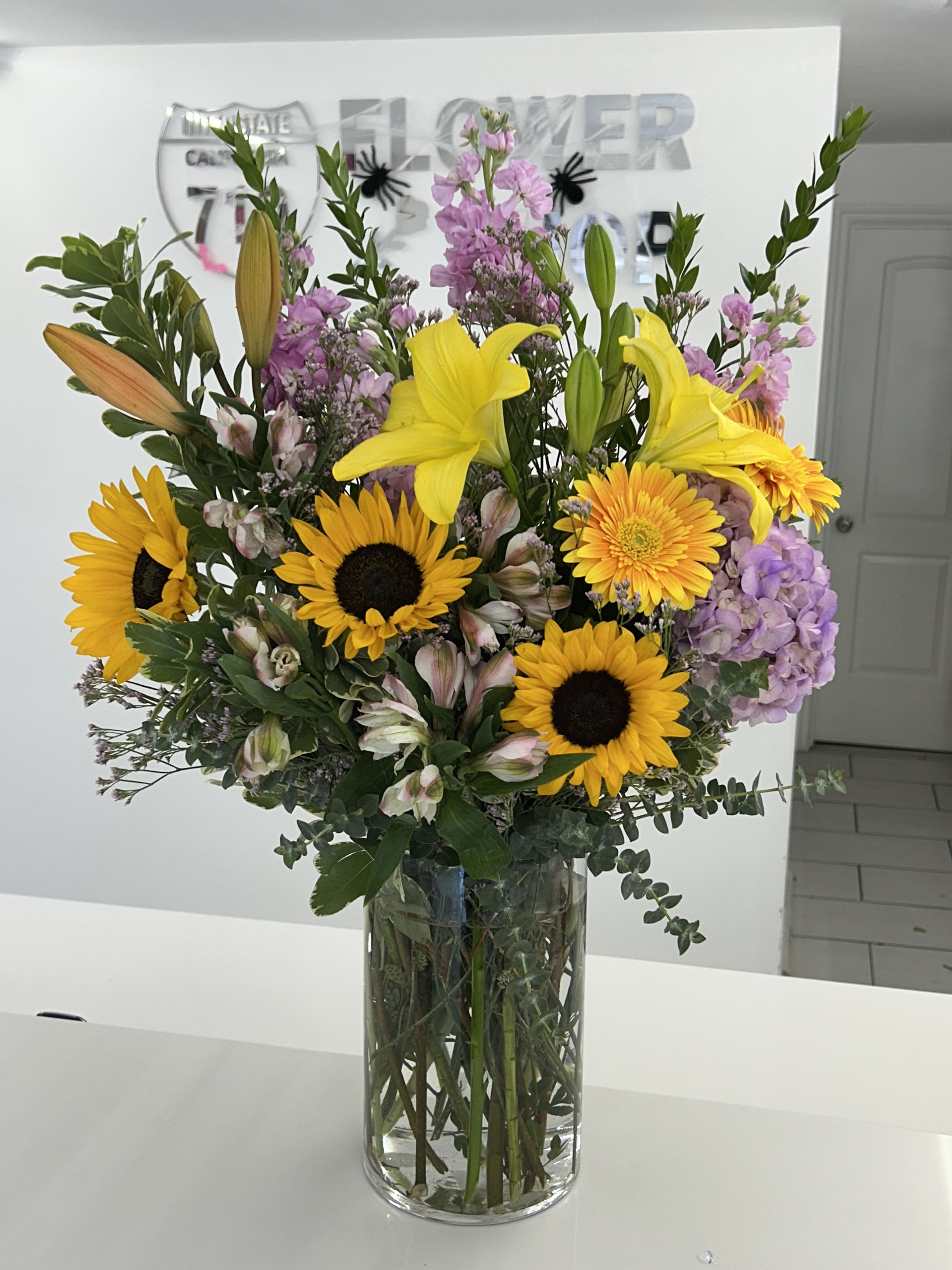 Bright Days Ahead - Like a breath of fresh air from a blooming summer field, this tall and cheerful bouquet features vibrant sunflowers, bright lilies, soft purple stock, and delicate alstroemeria. Arranged in a clear glass vase, it brings warmth, light, and joy to any room or celebration. (If some flowers and/or the vase are not available there will be minor substitutions.) 