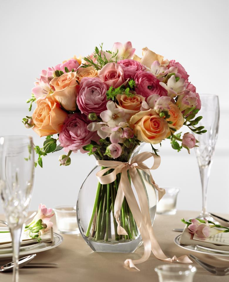 Pink and peach roses and ranunculus in a clear glass vase with a ribbon bow