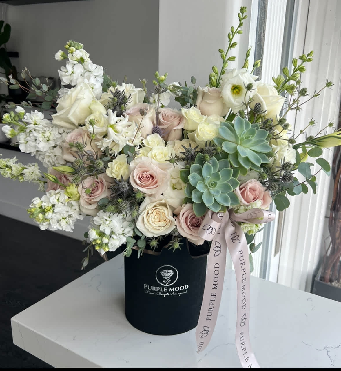 Natural Box - Container size - D6.75'x H7.5'  The Natural Box brings effortless beauty with soft neutral tones, fresh textures, and a hint of greenery. Styled in a sleek black hat box with ribbon, this arrangement is modern yet timeless — perfect for both gifting and home décor.  Key Features:     •    Styled in a matte black hat box for a modern finish     •    Accented with signature ribbon for elegance     •    Neutral, versatile color palette that suits any setting     •    A refined choice for birthdays, thank-yous, or stylish interiors