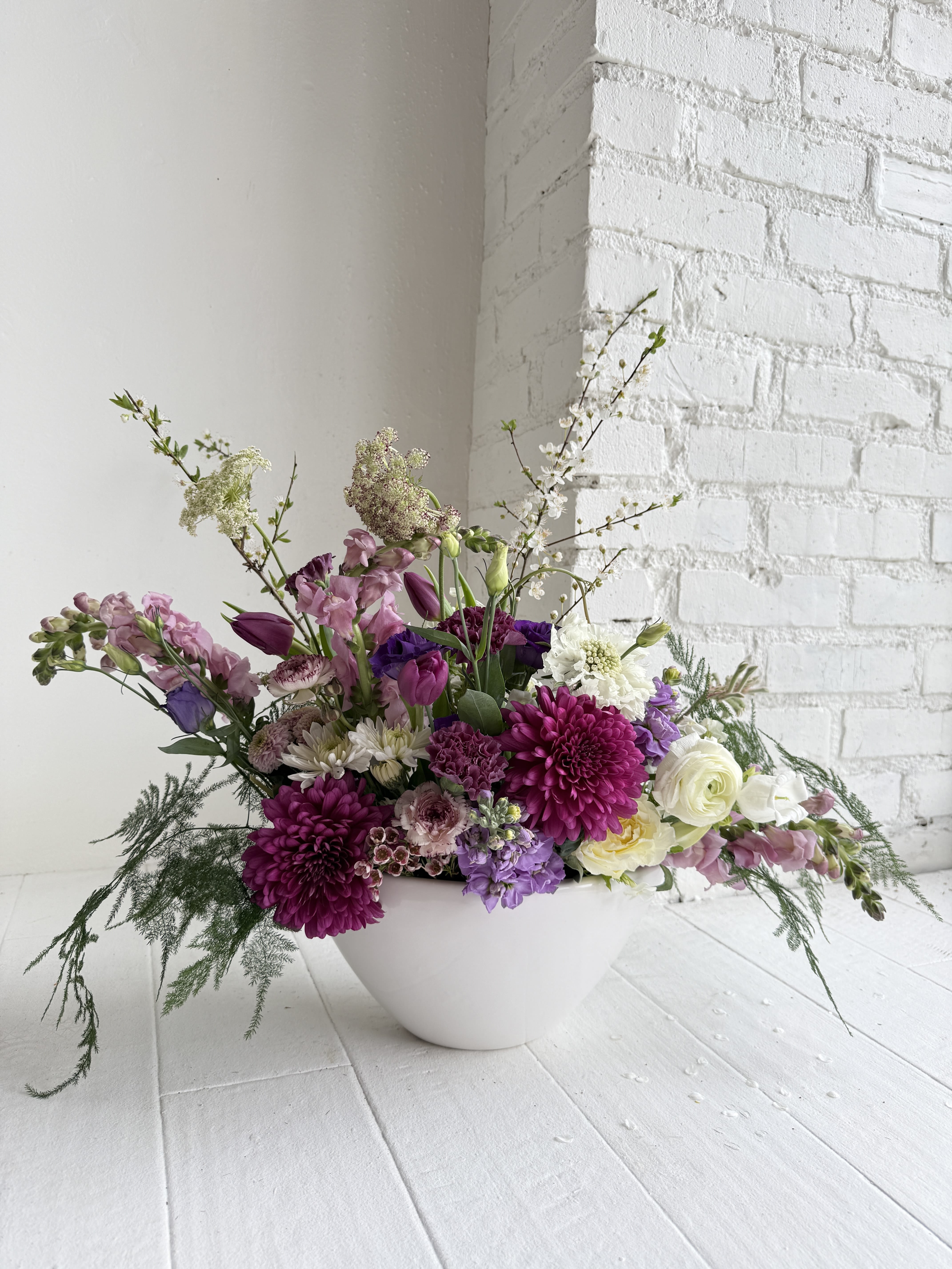 Violet - This simple but beautiful arrangement is composed with varying heights and textures. Inspired by our most requested color pallet; beautiful hues of purple and white in a white ceramic boat shaped vase.
