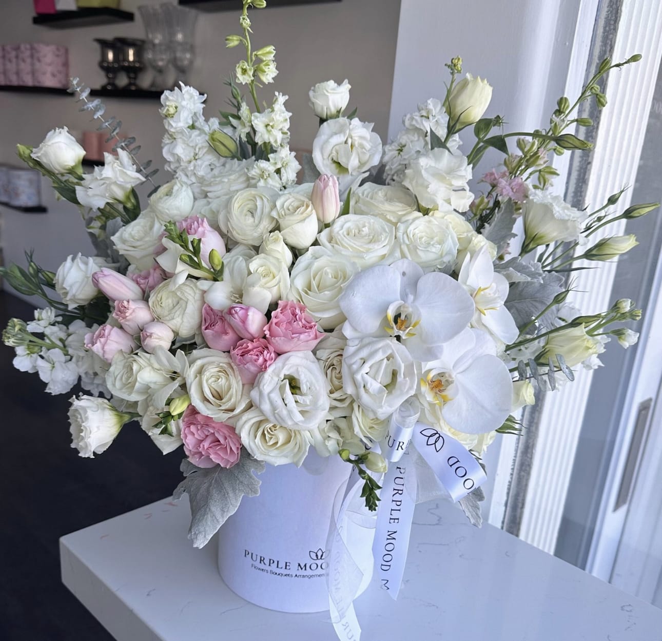 Perfect Anniversary Box - Standard container size - D6.75'x H7.5' Deluxe container size - D8.25'x H9.5' Premium container size - D10.5'x H12.25'  Celebrate love in its purest form with the Perfect Anniversary Box. A graceful mix of soft whites and blush pinks with elegant touches of orchids, this design radiates timeless romance. Refined and luminous, it’s the bouquet that says forever.      •    Elegant combination of white and blush tones     •    Luxurious orchids as a highlight     •    Presented in a signature hat box for a chic finish     •    Perfect for anniversaries, milestones, or classic romance gestures  Front side arrangement with lid in the back. Box includes wet floral foam inside, box doesn't require vase.