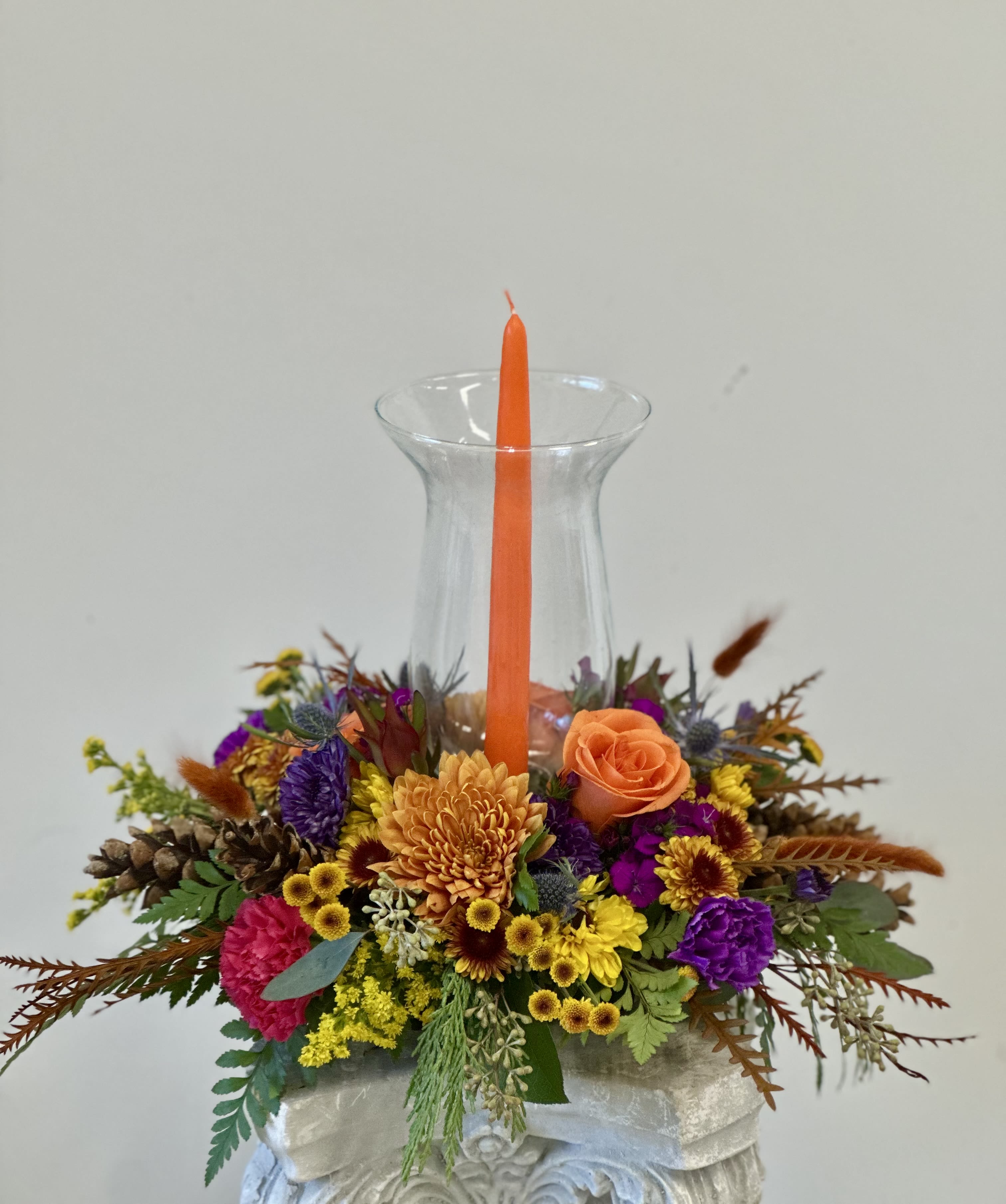 Harvest Sunset by Westford Florist Art by Veny - Elevate your seasonal decor with Harvest Sunset by Westford Florist Art by Veny. This stunning arrangement features a vibrant blend of bright fall-colored mums and poms, delicately paired with elegant roses, sweet william, and solidago. Accented by the rustic charm of pinecones and thistle, this floral masterpiece is topped with rich autumn foliage that captures the essence of fall. Housed in a gorgeous hurricane glass globe adorned with a tall candlelight, Harvest Sunset adds warmth and sophistication to any setting. Whether for your home or as a thoughtful gift, this exquisite centerpiece brings the beauty of autumn indoors while creating an inviting ambiance that enchants everyone who encounters it. Treat yourself or someone special to this captivating celebration of nature's bounty.