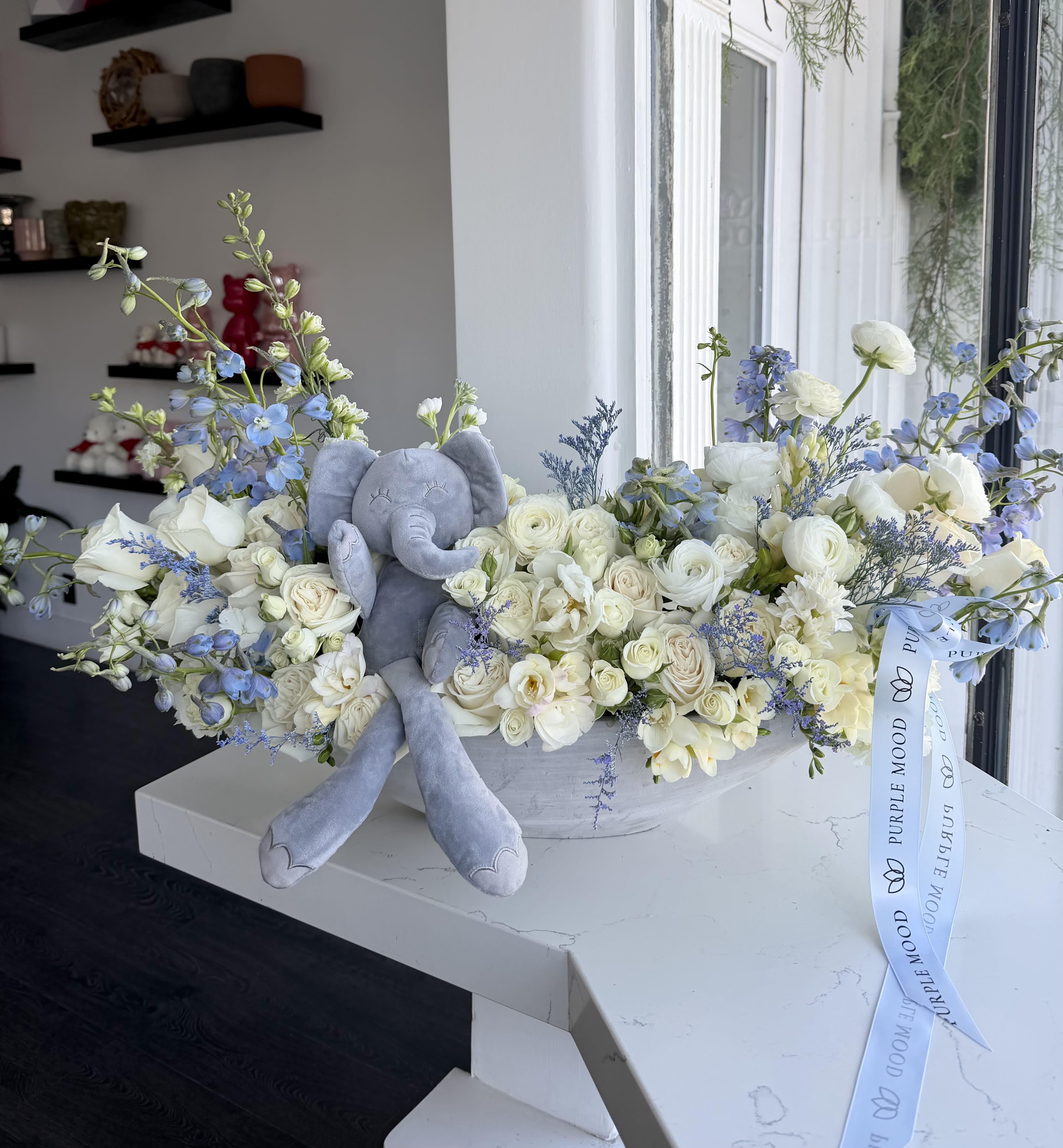 It's a Boy Vase - Celebrate the joy of a new beginning with the It’s A Boy Vase. A graceful arrangement in soft whites and gentle blues, accented with a plush elephant and elegant ribbons, makes this design a thoughtful gift for welcoming a baby boy. Both charming and sophisticated, it’s perfect for baby showers, hospital deliveries, or home celebrations.  Key Features:     •    Styled in a modern elongated vase with soft blue and white tones     •    Includes a plush elephant keepsake     •    Finished with signature ribbon for a polished touch     •    A heartwarming gift for baby showers, hospital visits, or welcoming baby home
