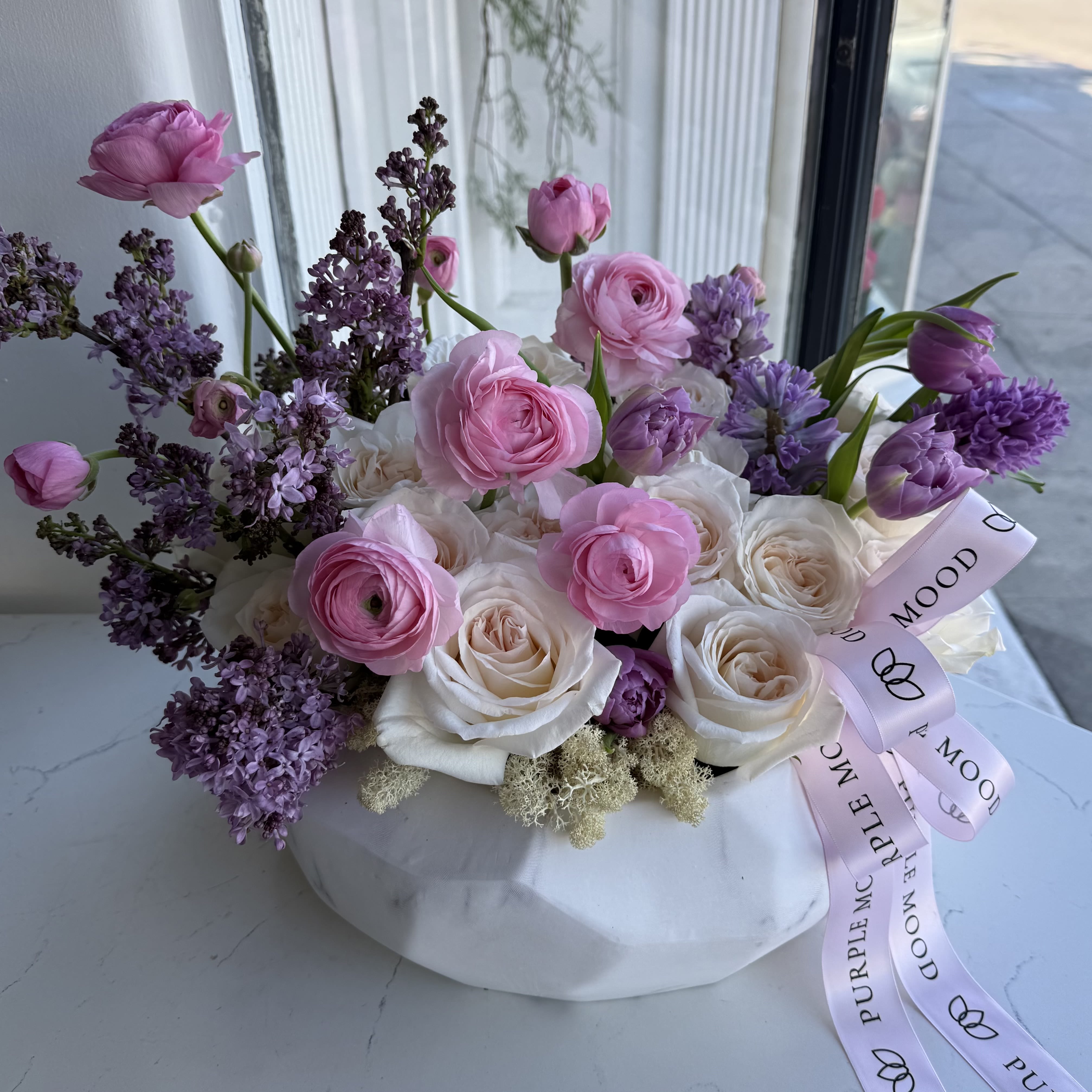 Lilac Vase - This elegant floral arrangement is presented in a sculptural ivory marble-inspired vase, designed to make a refined statement in any interior. A delicate blend of blush roses, soft ranunculus, and airy lilac blooms creates a harmonious palette of pastel pink and lavender tones.  The composition is thoughtfully balanced to achieve natural movement and depth, offering a look that feels both romantic and sophisticated.  Perfect for luxury interiors, celebrations, or as a signature gift, this piece embodies timeless beauty and modern femininity.  Each arrangement is handcrafted with precision and artistic attention to detail, ensuring that every creation is uniquely graceful.