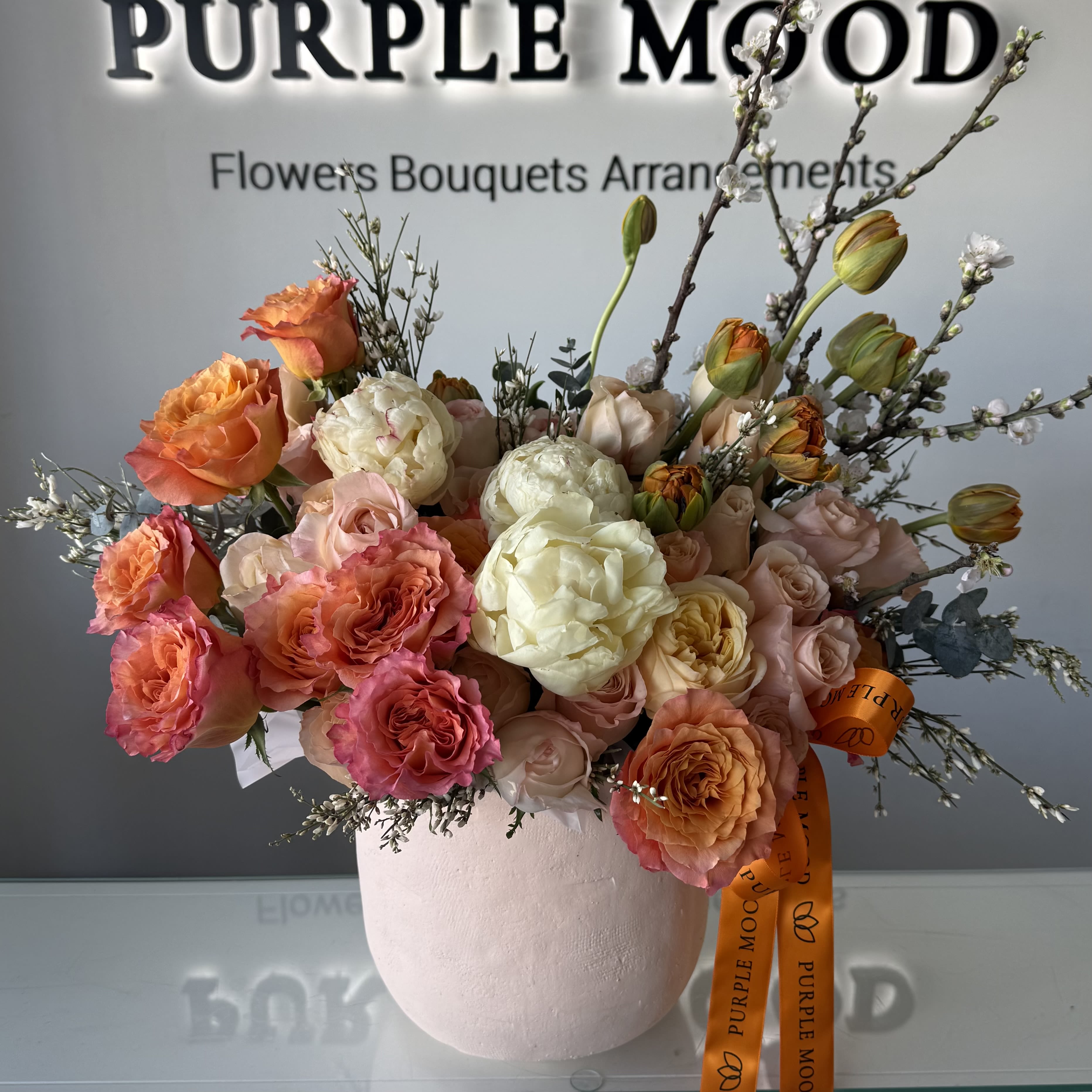 Sunset Crème Vase -  A refined arrangement featuring lush garden roses in creamy ivory, soft blush, and warm apricot tones, accented with delicate seasonal blooms and natural branches. Designed to evoke the glow of a sunset, this vase arrangement brings warmth, elegance, and timeless beauty to any space.