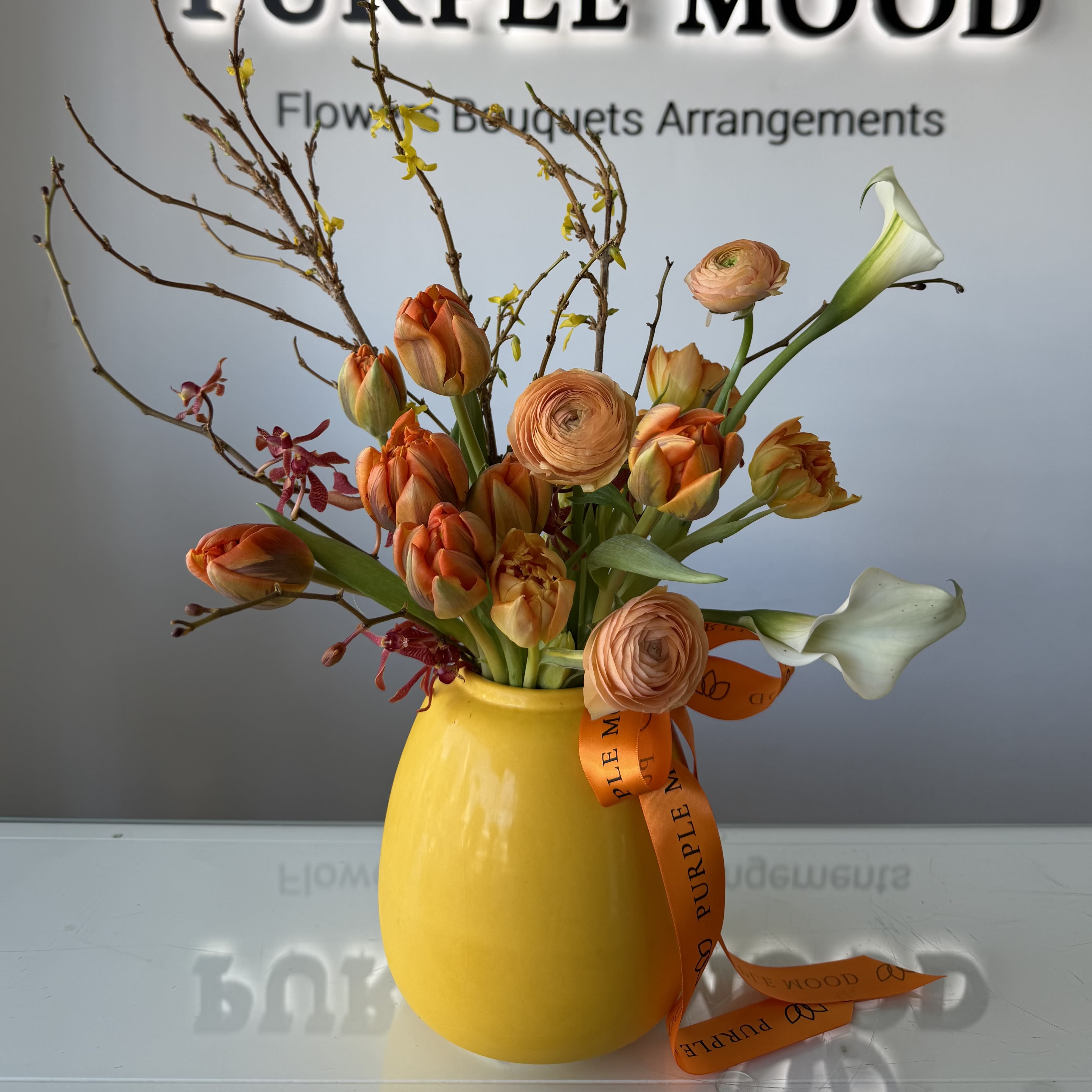 Orange Glow Vase - A charming small arrangement designed to bring warmth and light into any space. Featuring glowing orange tulips, delicate ranunculus, elegant white calla lilies, and seasonal flowering branches, this compact design is arranged in our signature vase for a refined, modern look.  Perfect for desks, coffee tables, or as a thoughtful gesture when you want something petite yet full of personality.