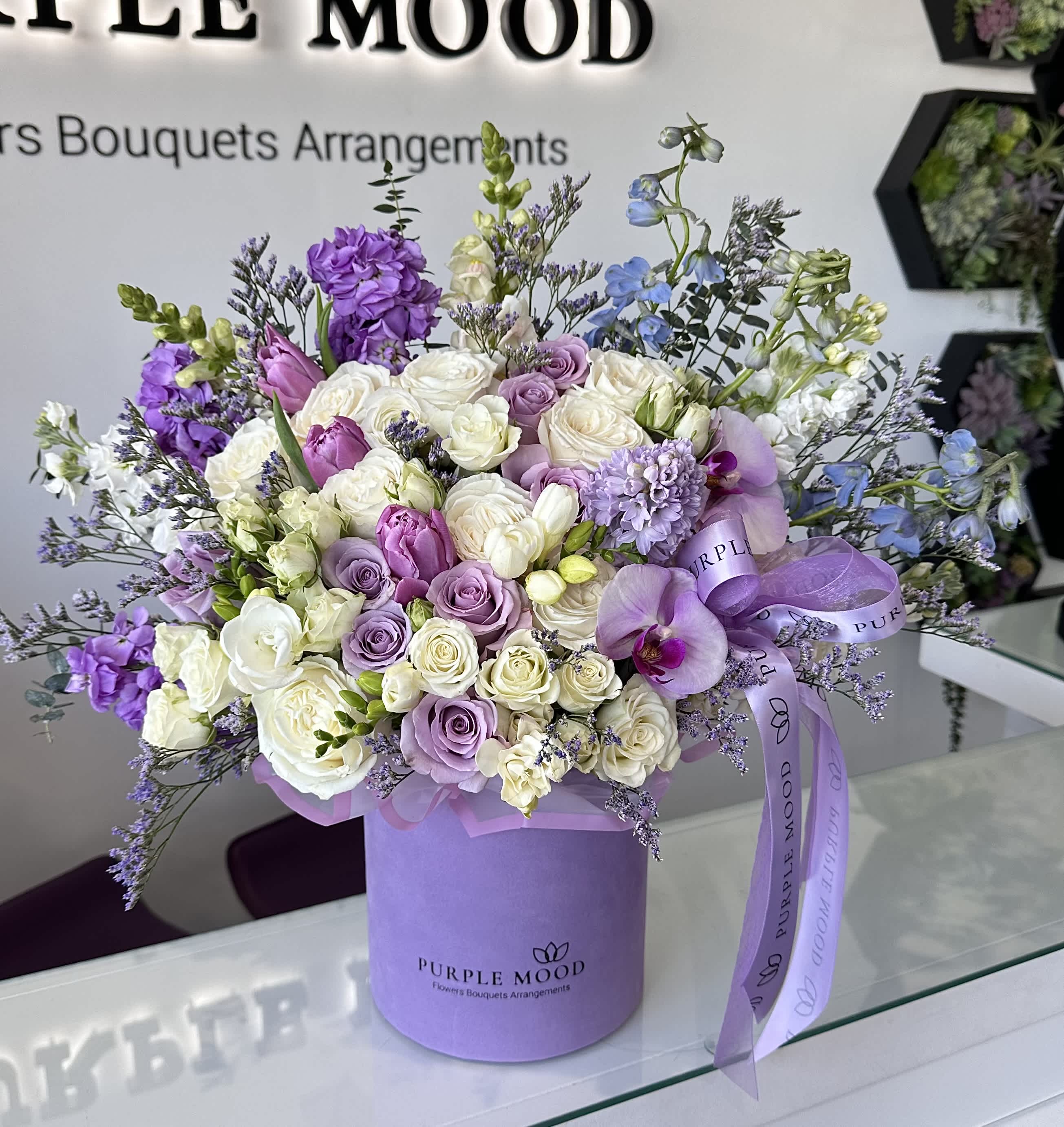 Dreamy Garden  - Lavender box with white and lavender flowers.   Standard - Container size D6.75'x H7.5'  Deluxe - Container size D8.25'x H9.5'  Premium - Container size D10.5'x H12.25'  An enchanting arrangement that captures the beauty of a fairytale garden. Featuring a lush mix of roses, lisianthus, orchids, stock flowers, and seasonal greenery, this bouquet brings together soft shades of purple, cream, and blush. Elegant and romantic, it’s the perfect centerpiece for weddings, anniversaries, or any moment that deserves to feel unforgettable.    •    Premium blooms: roses, lisianthus, orchids, stock, and seasonal fillers     •    Color palette: soft purples, creams, blush pinks, and ivory