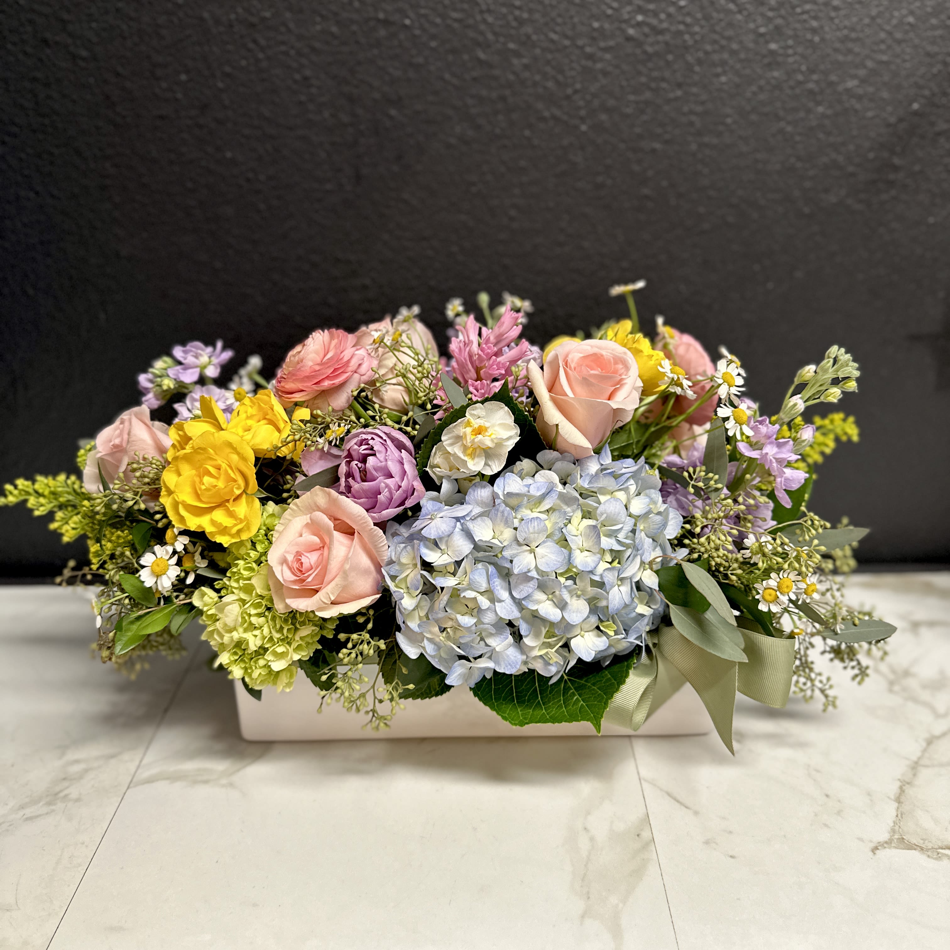 Spring Pastel Centerpiece - A designer's choice Spring Pastel garden mix of the season's freshest blossoms designed in a low rectangular vase. The varieties will vary, the color tones can include soft pinks, peach, lavender, butter yellows, soft blues, tones of green and accents of white.  This design is intentionally low so your seated guests can easily see over the flowers. Arranged in our 4" x 4" x 12" white ceramic centerpiece vase. 