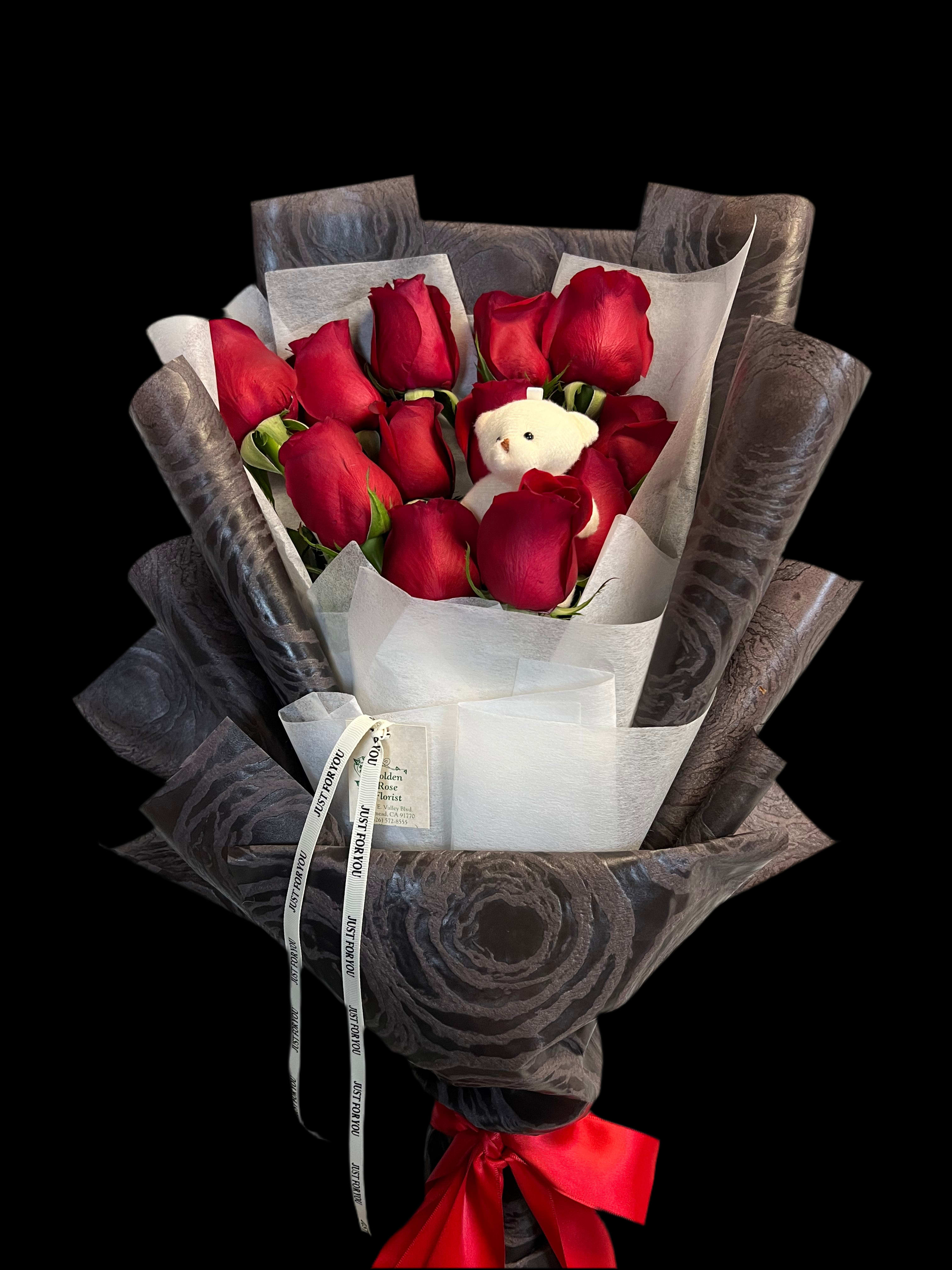 Hand-tied bouquet of red roses wrapped in dark paper with a small white teddy bear.