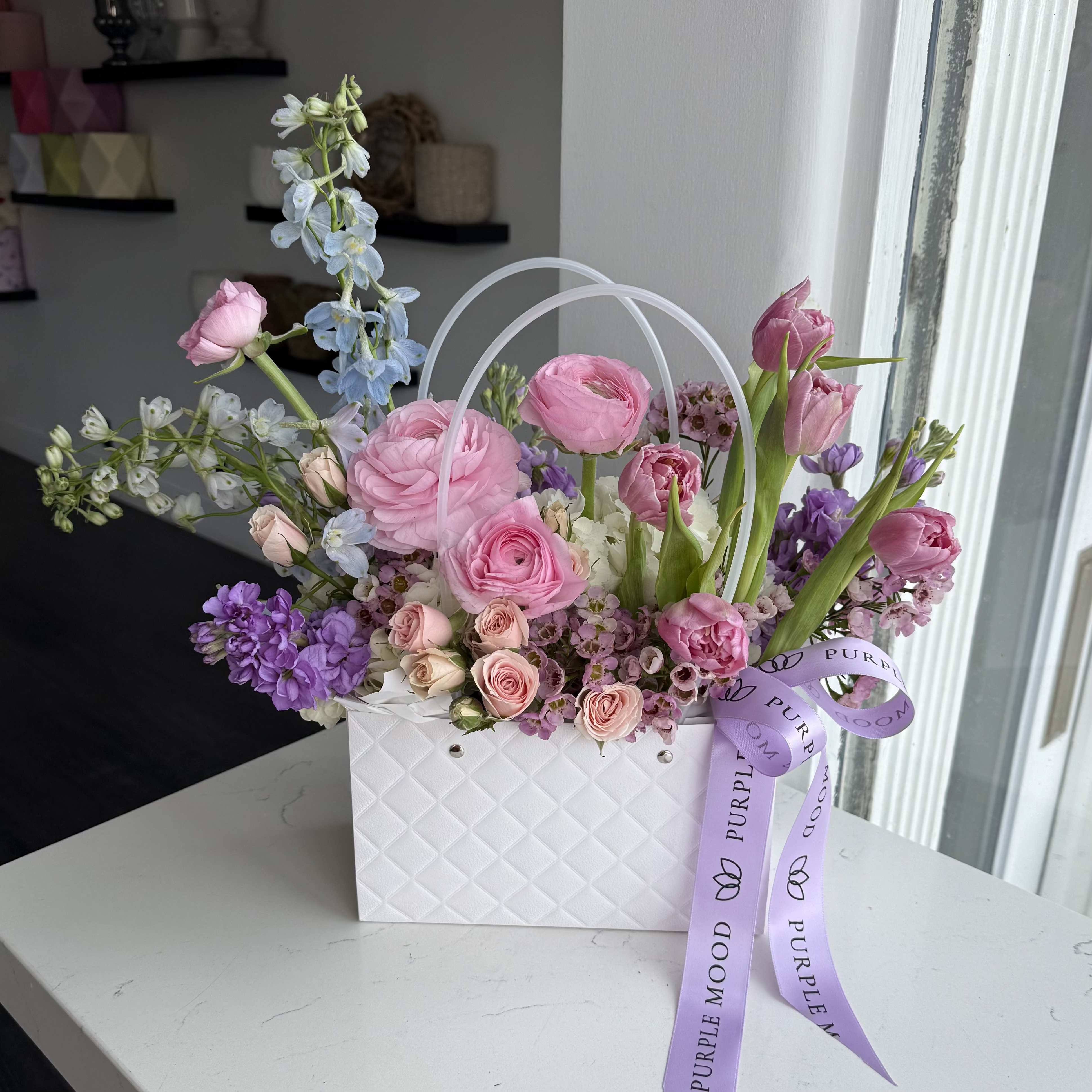 Mini Bag White - (Flowers can be different)  Elegant and sweet, the Mini White Bag is a charming arrangement of pink and purple blooms presented in a quilted white bag with handle. Compact yet eye-catching, it’s the perfect little gift to brighten someone’s day with a touch of beauty and grace.  Key Features:     •    Accented with a signature ribbon for a polished finish     •    Compact size, ideal for desks, bedside tables, or thoughtful surprises     •    A lovely and versatile gift for birthdays, thank-yous, or just because