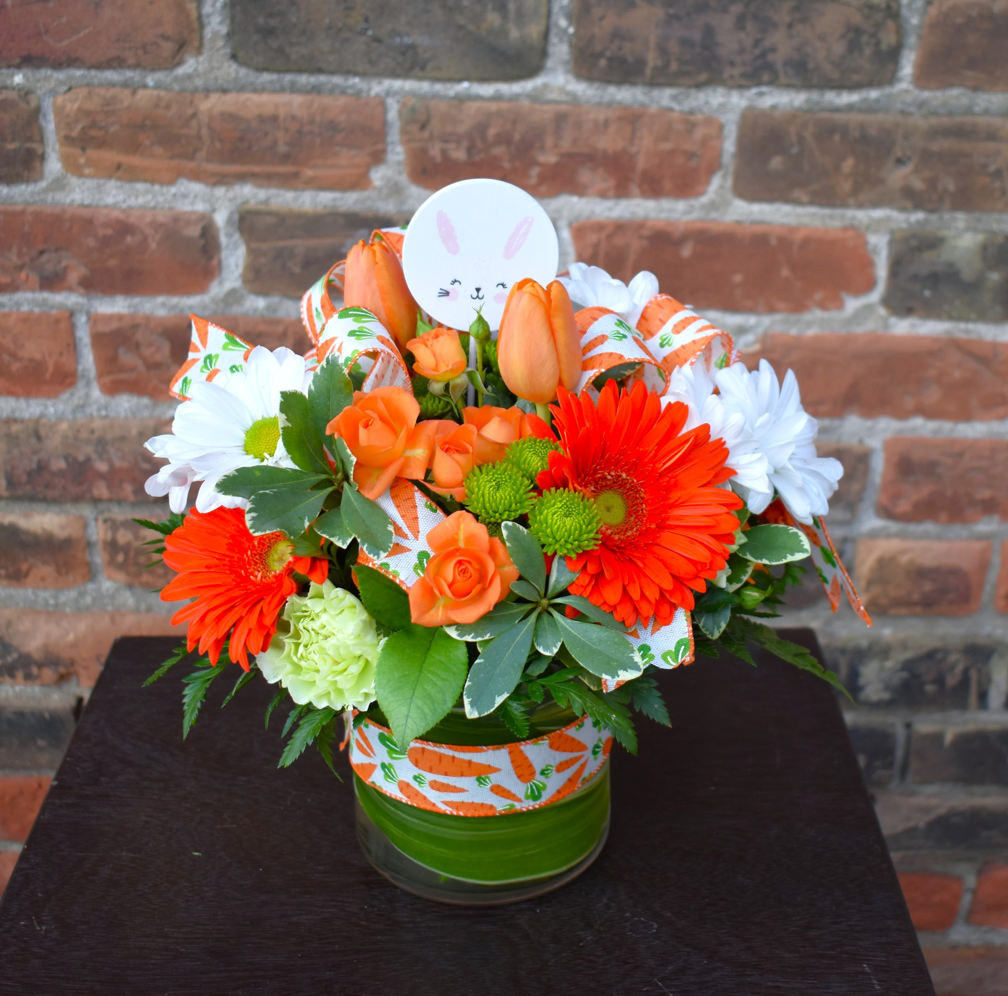 Carrot Patch - A bright and fun arrangement featuring white daisies, green button mums and carnations, orange gerber daisies, spray roses and tulips, lush greens accented with carrot-ribbon and a cute bunny pick...the perfect design to celebrate spring!  Approx Size: 1ft X 1ft