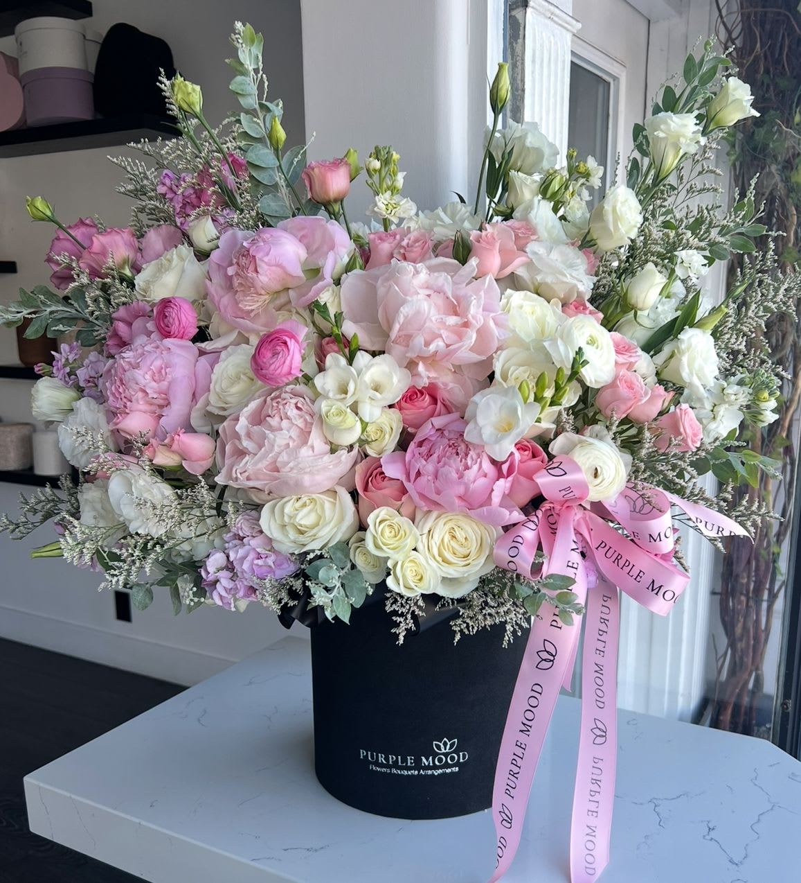 Peony BOOM - Standard container size D8.25'x H9.5' Deluxe container size - D10.5'x H12.25'  A statement piece overflowing with romance and luxury — the Peony BOOM arrangement is pure elegance in bloom. Bursting with layers of soft pinks and creams, it’s designed to impress and delight on the grandest scale.      •     Lush, voluminous design for a true show-stopper look     •     Perfect for anniversaries, weddings, or unforgettable surprises     •     Elegant presentation in a signature box with satin ribbon  Front side arrangement with lid in the back. Box includes wet floral foam inside, box doesn't require vase.