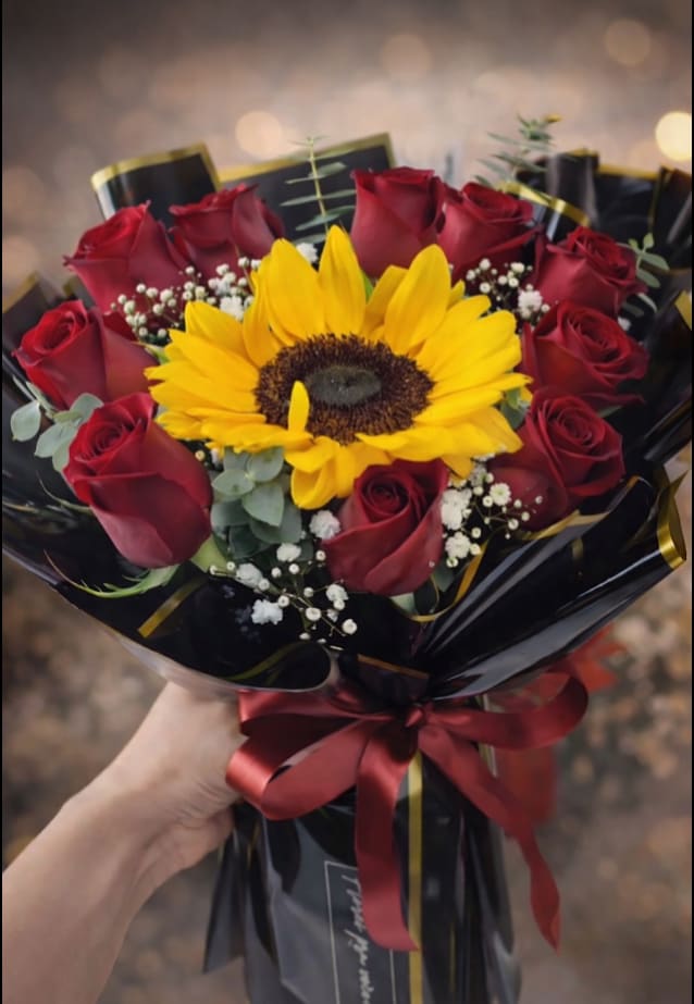 Golden Love Bouquet - A single oversized sunflower crowns this hand-tied flower arrangement, its golden face framed by a ring of deep red roses-the perfect birthday bouquet or anniversary roses with a modern, dramatic twist. The red-and-yellow palette reads both celebratory and romantic, so the person you're thinking of will feel noticed and cherished whether you're saying "happy birthday," "congratulations," or "I love you."  Small white filler blooms are scattered through the design to soften the contrast and add delicate texture, while the stems are gathered in glossy black cellophane wrap edged in gold and secured with a wide red satin ribbon and label tag. Finished in black plastic bouquet wrap for easy handling, this arrangement arrives gift-ready and polished; it's an energetic, contemporary choice for anyone who loves bold color and striking presentation.