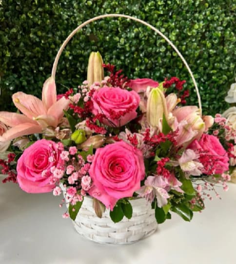 Love in a Basket - A compact cluster of vivid pink roses and pale pink lilies arranged in a white wicker basket creates an immediate impression of cheerful romance-perfect as a birthday bouquet, anniversary gift, or a spontaneous "thinking of you" surprise. This flower arrangement for celebrations and affectionate occasions makes the recipient feel seen and adored with its soft palette of pinks, whites, and touches of red.  The arrangement pairs roses, lilies, alstroemeria, and delicate pink filler blooms with sprigs of deeper red flowers for contrast, all gathered low in a white woven basket with an arching handle. The basket's airy frame and the mix of textures-lush rose petals, graceful lily blooms, and dainty alstroemeria-give the piece a charming, garden-fresh look that fits on a table or bedside. Sizes and care details are provided at checkout; stems are arranged for easy display and should be kept in fresh water to extend bloom life.
