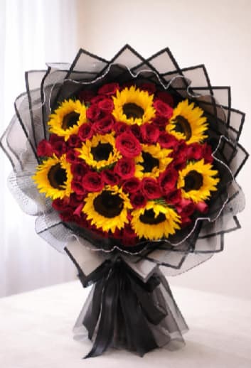 lden Queen Bouquet - Sunflowers and deep red roses combine for a bold, celebratory statement in this large hand-tied flower arrangement-perfect as a birthday bouquet, anniversary roses, or a romantic everyday gift. The bright yellow sunflower faces and velvety red rose clusters are arranged in a tight, rounded dome so the recipient will be greeted with an immediate, joyful burst of color and contrast the moment they open it.  Tightly grouped blooms are framed with layered black mesh and translucent paper wrap that fan out behind the flowers, creating a dramatic, graphic silhouette. Stems are gathered with a wide black ribbon for a polished, contemporary finish; the mix of warm petals and structured dark accents gives the bouquet strong visual impact and modern flair.  Choose this arrangement when you want someone to feel noticed and celebrated-the vivid palette reads as both festive and intimate, an effortless way to say "I'm thinking of you." This large hand-tied design arrives ready to display; trim stems and place in fresh water to extend vase life.