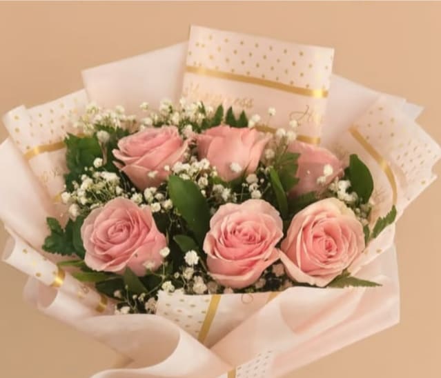 Subtle Elegance - Soft pink roses gathered in a close, romantic cluster create the first impression of this flower arrangement - a delicate birthday bouquet or a thoughtful way to say "thank you" or "I'm thinking of you." Tiny white filler blooms peek between the roses, softening the silhouette and giving the bouquet a light, airy texture that will make the recipient feel genuinely seen and cherished.  Wrapped in layered pink and white paper patterned with gold dots and lines, the presentation feels polished and celebratory without being overstated. The mix of rose petals and petite white accents offers gentle contrast in both color and texture; the finished size makes a lovely hand-held bouquet that suits intimate moments - anniversaries, romantic gestures, or lifting someone's day. For best longevity, keep stems in fresh water, recut stems before placing in a vase, and display away from direct heat and drafts to prolong bloom life.