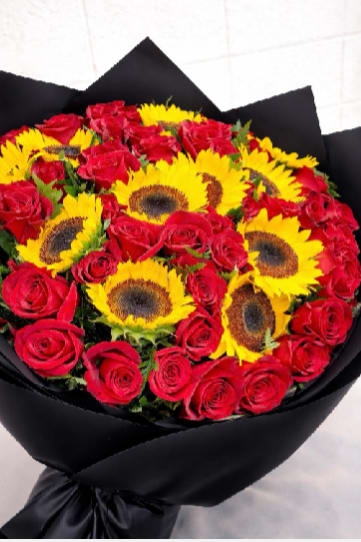 Sunflower &amp; Roses Luxury - A radiant cluster of bright sunflowers paired with deep red roses makes this hand-tied flower arrangement an unforgettable choice for an anniversary bouquet or bold birthday surprise. The contrast of golden yellow and velvety red reads as both celebratory and romantic, so the person you're thinking of will feel seen and celebrated the moment they open it.  The round, tightly gathered design emphasizes striking color blocks of sunflowers and roses, with lush green foliage peeking through for depth. Wrapped in smooth black paper and tied with a black ribbon, the modern, luxurious presentation adds drama and polish-perfect for someone who loves contemporary style. The compact, hand-tied shape is easy to display in a vase and keeps the focus on the vivid blooms.  Ideal for romantic gestures, milestone celebrations, or anyone who responds to bold color, this arrangement sends a confident, joyful message. To keep the bouquet looking fresh, recut stems and change the water every couple of days; arranging in a shallow vase suits the rounded shape best.