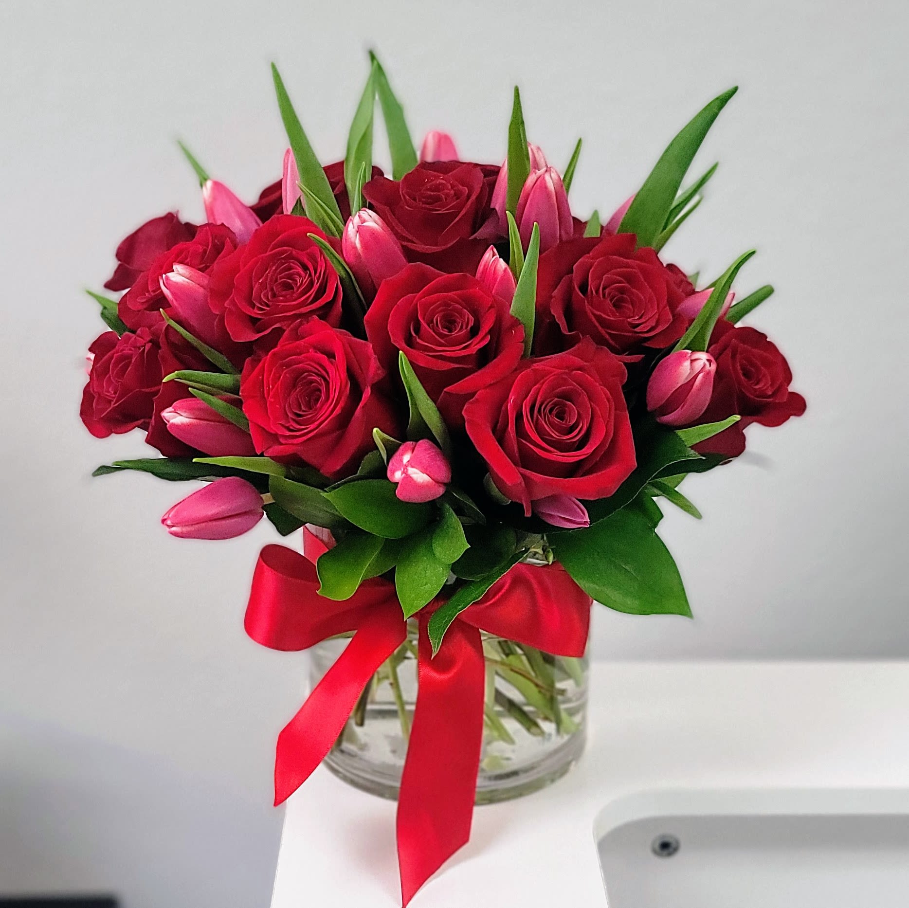 Speak Your Heart Bouquet - Express your love in full bloom with this stunning combination of red roses and delicate tulips. Fragrant crimson roses symbolize passionate love, while pink tulips represent enduring affection and devotion. This beautiful bouquet is sure to leave a lasting impression.  Flowers: 10 Red Roses 20 Pink and Red Tulips  Hand crafted and finished with a satin bow Vase: Delivered in a glass cylinder vase  Dimensions: Approximately 14" W x 13" H Orientation: All-Around