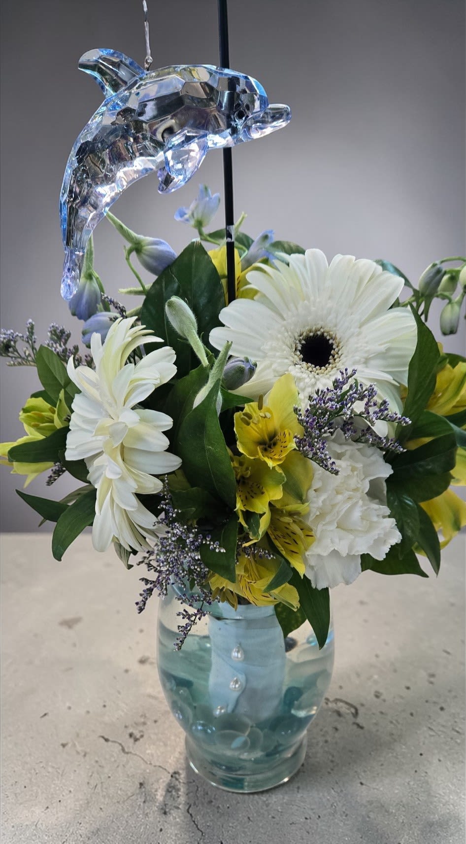 Ocean Whisper - A coastal-inspired arrangement in a clear vase with aqua accents, featuring white gerbera daisies, carnations, yellow alstroemeria, and soft purple fillers. Topped with a sparkling crystal dolphin, it feels fresh, airy, and seaside-inspired.