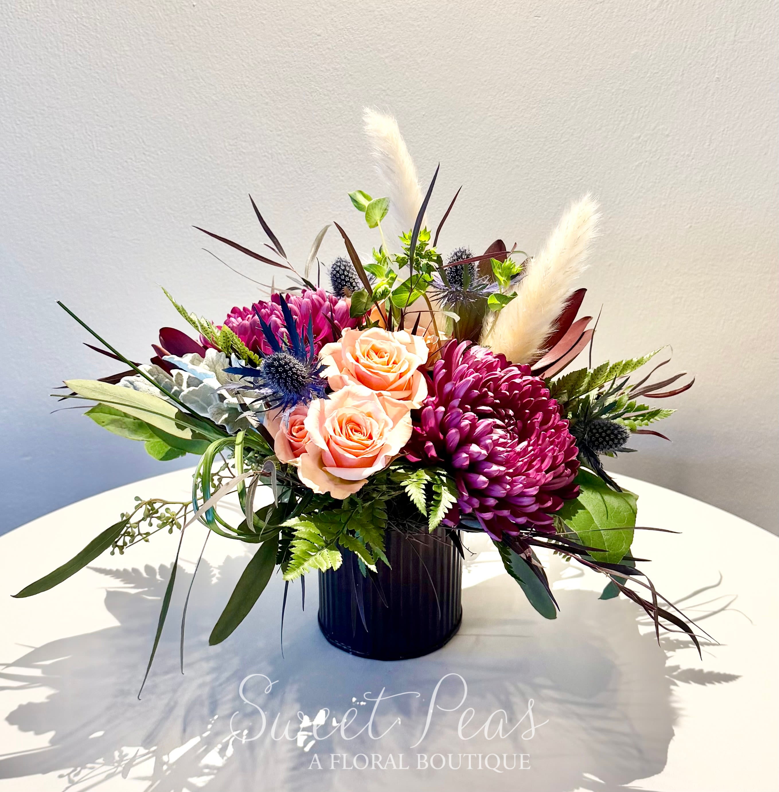 Nature Walk  - Rich and textural, 'Nature Walk' features soft peach spray roses paired with deep plum mums, sculptural blue thistle, and airy seasonal accents. Designed with flowing greenery and wisps of dried elements for movement and contrast, this arrangement is artfully styled in a sleek black ceramic vessel. Perfect for gifting, sophisticated celebrations, or anyone who loves modern floral design with depth and character.