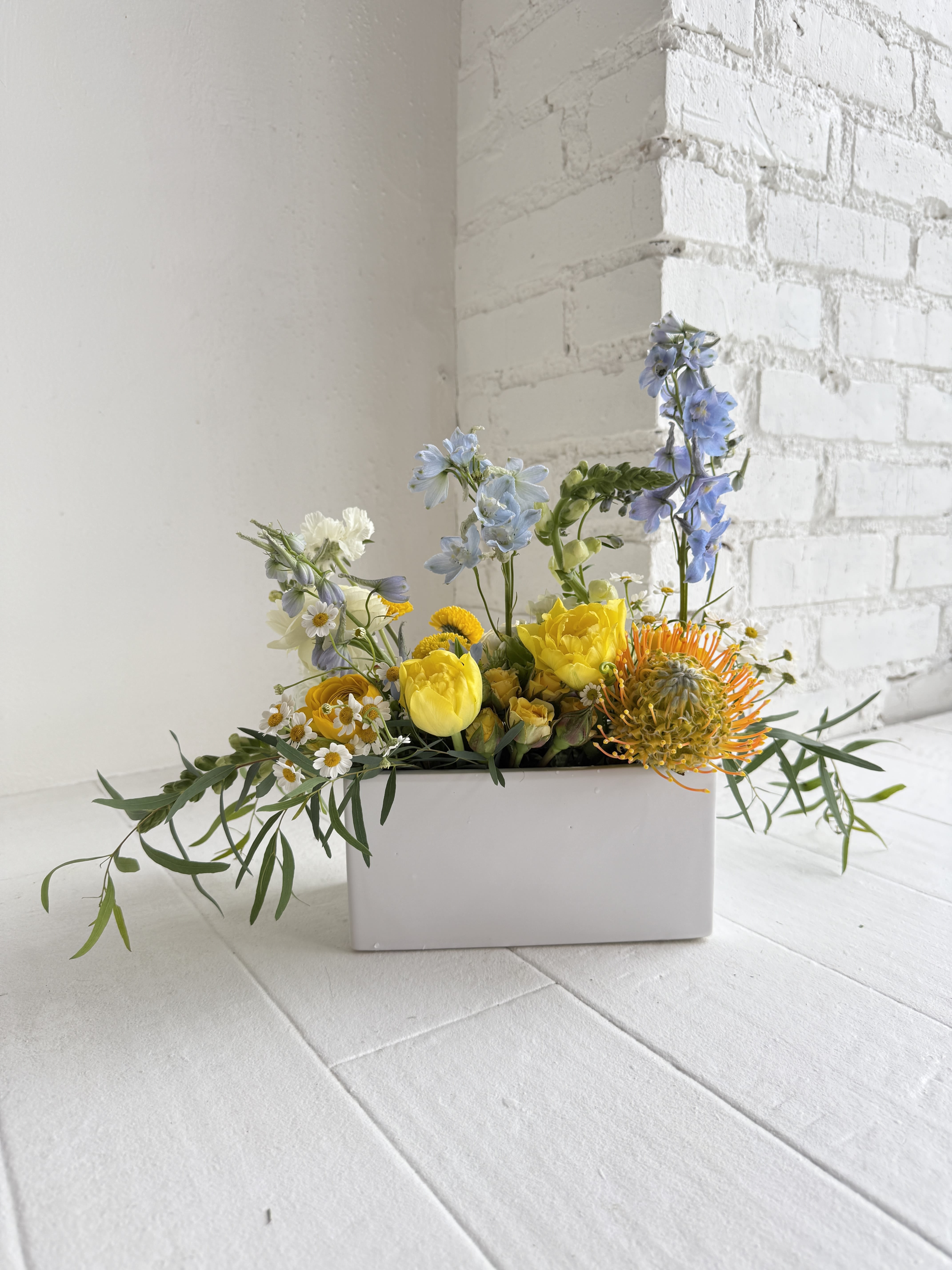 Clara - A pretty mix of blues, yellows and whites in a low, white, rectangular ceramic vase. Please note that some flower substitutions may need to be made, but the arrangement will have the overall same look and feel.