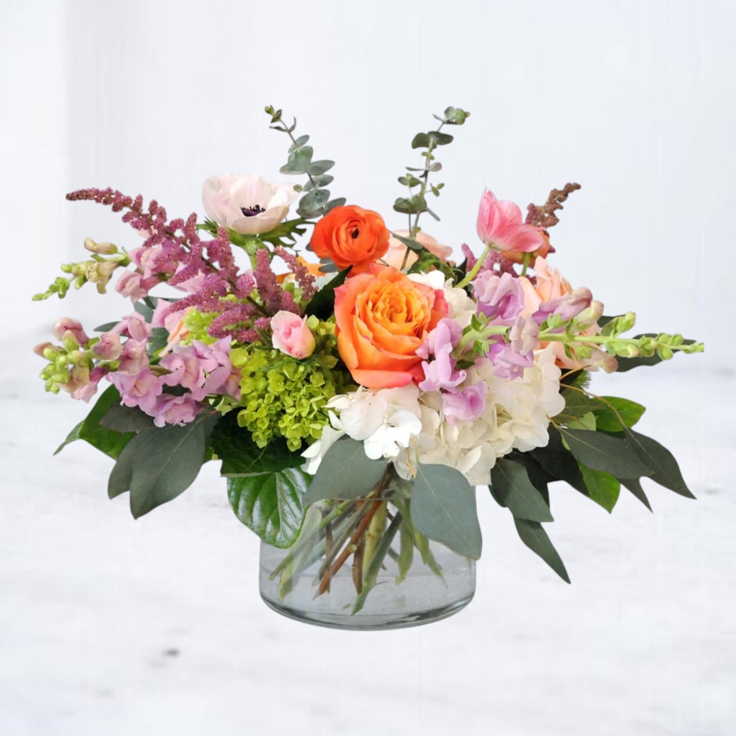 Bright Mix - A spreading cluster of bright blooms gives this mixed-color flower arrangement an immediate burst of sunshine-perfect as a birthday bouquet or a cheerful everyday gift for the person you're thinking of. Soft pink ranunculus and lavender-toned blooms mingle with white and green hydrangeas to create a joyful, uplifting palette that reads as both celebratory and comfortably modern.  Low and abundant, the design sits in a clear glass cylinder vase that keeps the look fresh and contemporary while showcasing the dense, textural base of hydrangea. Snapdragons, and a few airy accent stems add graceful height and movement; spray roses, anemone, and other small flowers provide delicate detail throughout. Sized to brighten a kitchen table, desk, or bedside, it arrives ready to enjoy and brings a noticeable burst of color and warmth to any celebration.