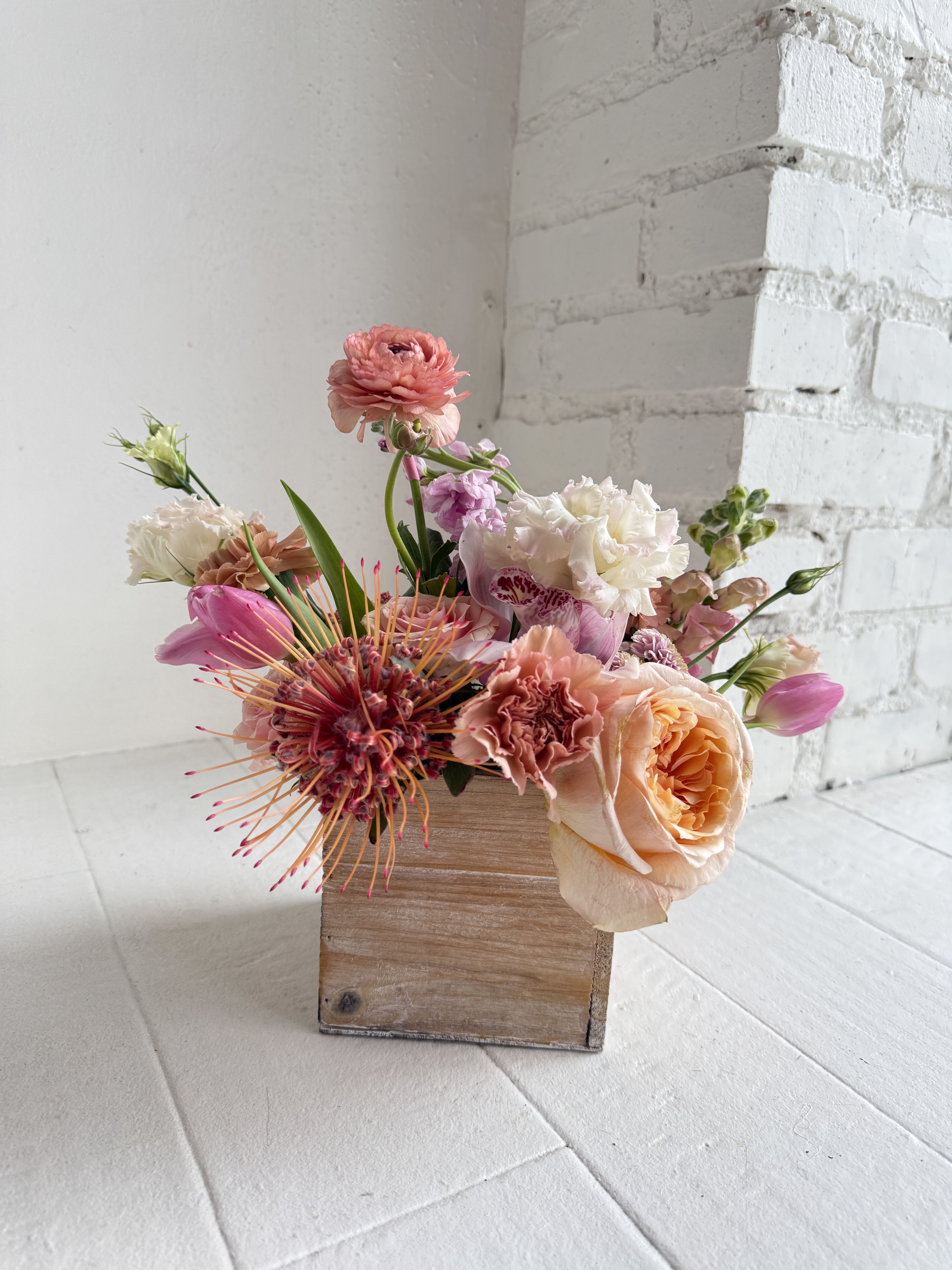 Zoe - Zoe. This arrangement is stunning and sure to please. Flowers to the max! We take a handcrated wooden crate and artfully arrange our favorite seasonal blooms in saturated shades of pink and peach with pops of white flowers. An abundance and focus of in season flowers will be used. We favor roses, stock, snapdragon, ranunculus, peonies, lisianthus, tulips, anemones, protea, wax flower.