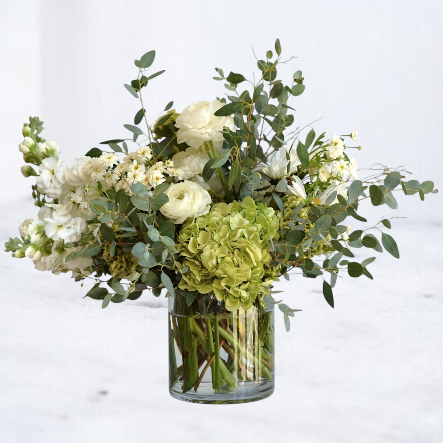Blissed - Soft, serene, and effortlessly refined, Blissed features lush white hydrangea paired with creamy ivory roses, layered with fresh eucalyptus and delicate seasonal greenery. Designed in a clear glass cylinder vase, this arrangement balances fullness with natural movement for a look that feels timeless and calming. Perfect for sympathy gestures, thank-you gifts, housewarmings, or moments when quiet elegance speaks louder than words.  Designed with premium seasonal blooms. Flower varieties may vary slightly to ensure the freshest, most beautiful arrangement.
