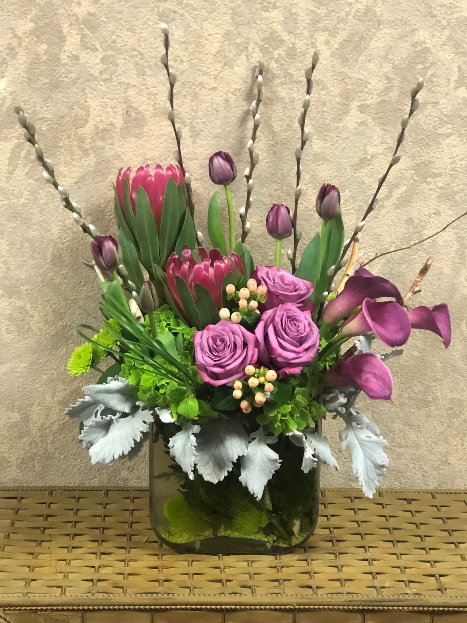Purple Elegance - Type of Flowers: Purple Protea, Purple Tulips, Lavender Roses, Purple Mini Calla Lillies, Pussy Willow, Green Hydrangeas, Peach Hypericum Berries, Branches, and Dusty Miller in a glass vase. Availability: All year round Substitute Available: Yes Design View: All Facing View Photo shown: Regular