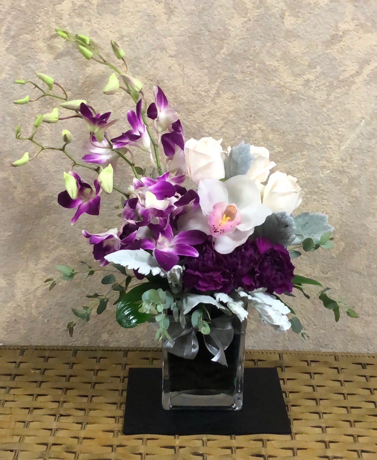 Orchids for the Best - Type of Flowers: White Cymbidium Orchids, Purple Phalaenopsis Orchids, White Roses, Purple Carnations and Dusty Miller in a square vase with wrap and bow. Availability: All year round Substitute Available: Yes Design View: All Facing View Photo shown: Regular 