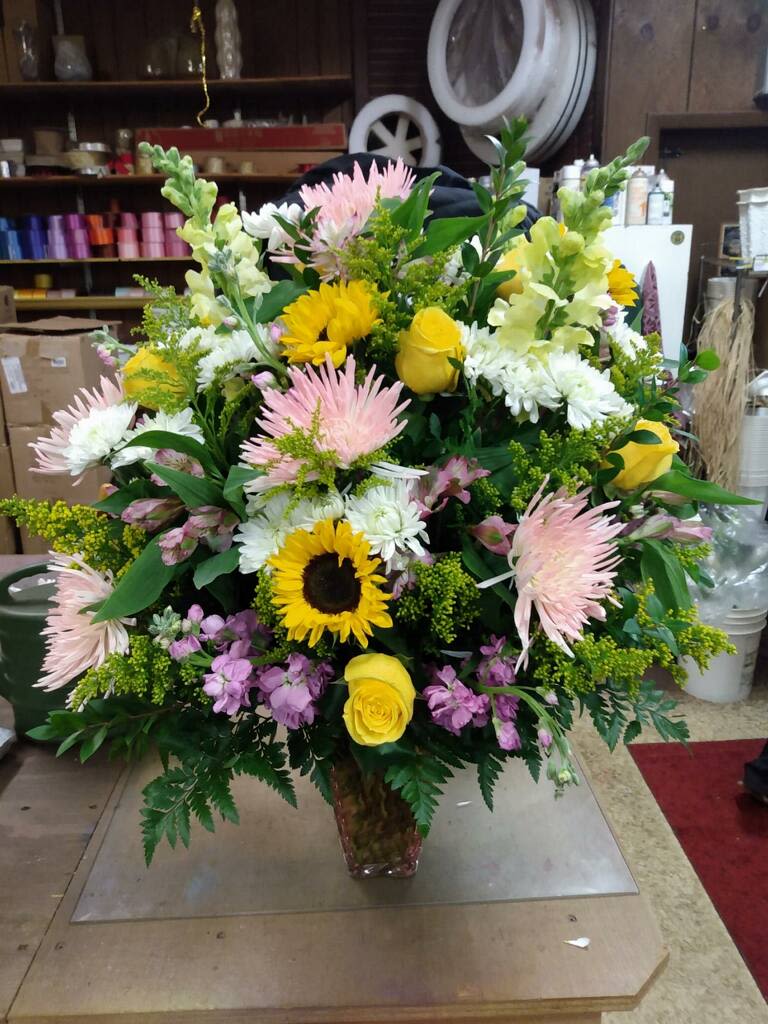 6304 Sympathy Fresh - One sided sympathy fresh includes, snap dragons, yellow roses, solidago, purple alstromaria, sunflowers, white cushion mums, spider mums greens in a versitile container.  (substitutions will be made to value upon availability)