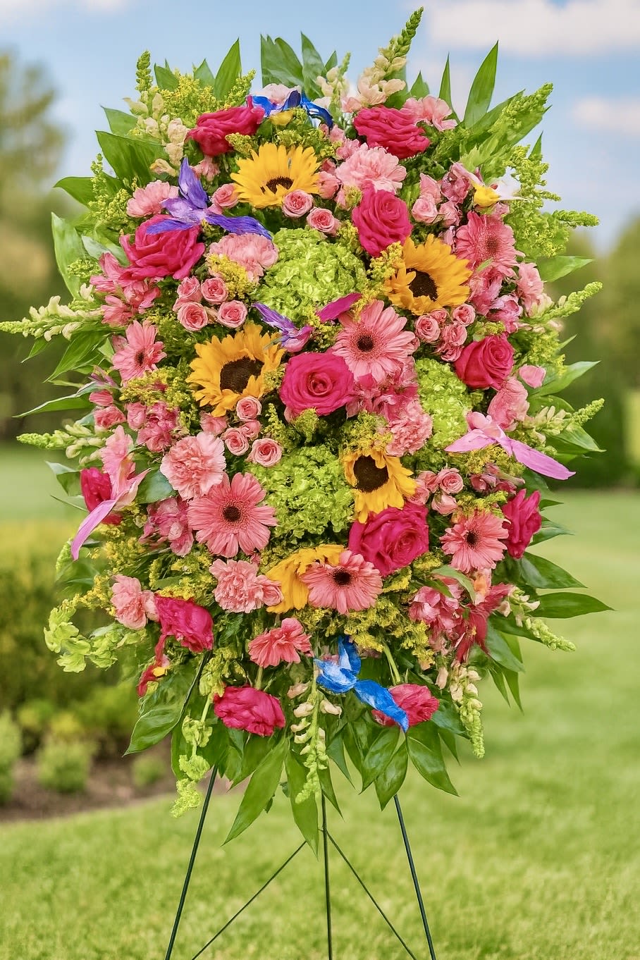 Tall standing spray of pink roses and gerberas with yellow sunflowers on a metal easel outdoors