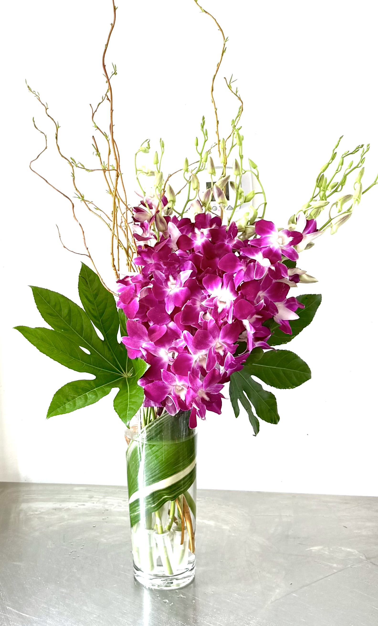 Orchid Embrace - Immerse yourself in the captivating elegance of this stunning arrangement, which features a generous bunch of purple and white Dendrobium orchids. These delicate, star-shaped blooms are gathered in a simple, clear glass vase. The design is enhanced by unique tropical foliage, which is delicately folded as if to lock the flowers together in a sweet embrace, and accented with curly willow branches. This simple yet elegant display of natural beauty is perfect for any occasion. 