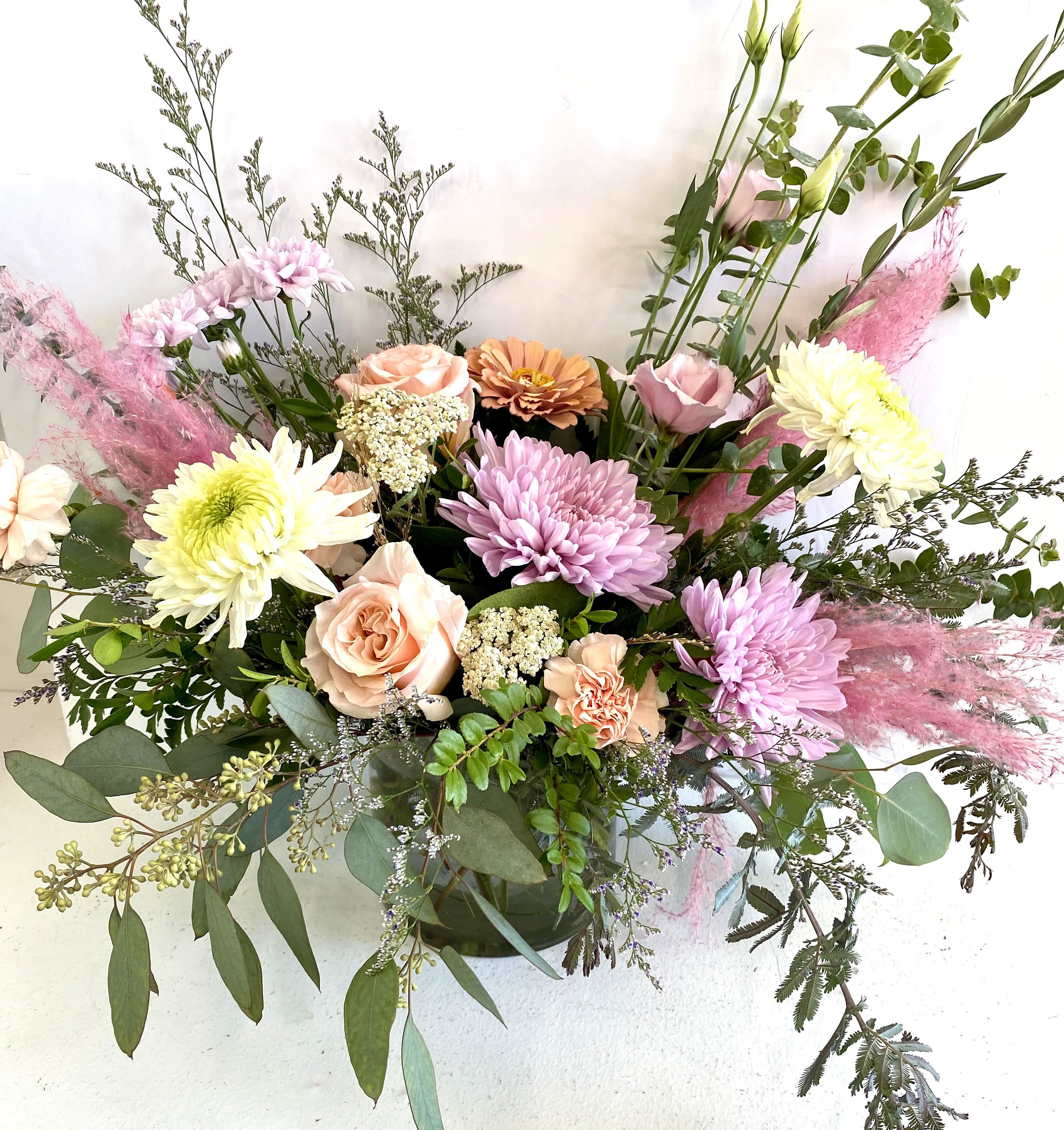 Floral Whimsy - Delicate yet vibrant, the Floral Whimsy arrangement captures the beauty of soft pastel tones in full bloom. This enchanting arrangement features elegant roses in peach and blush, complemented by bright chrysanthemums and soft carnations. Sprigs of eucalyptus and lush greenery add texture, while the subtle pink pampas grass creates an ethereal touch of whimsy. Perfect for a romantic gesture, a warm celebration, or as a thoughtful gift to brighten someone's day, this bouquet is designed to evoke calm and joy in every setting. Arranged in a chic vase, it's a stunning piece that effortlessly elevates any space.