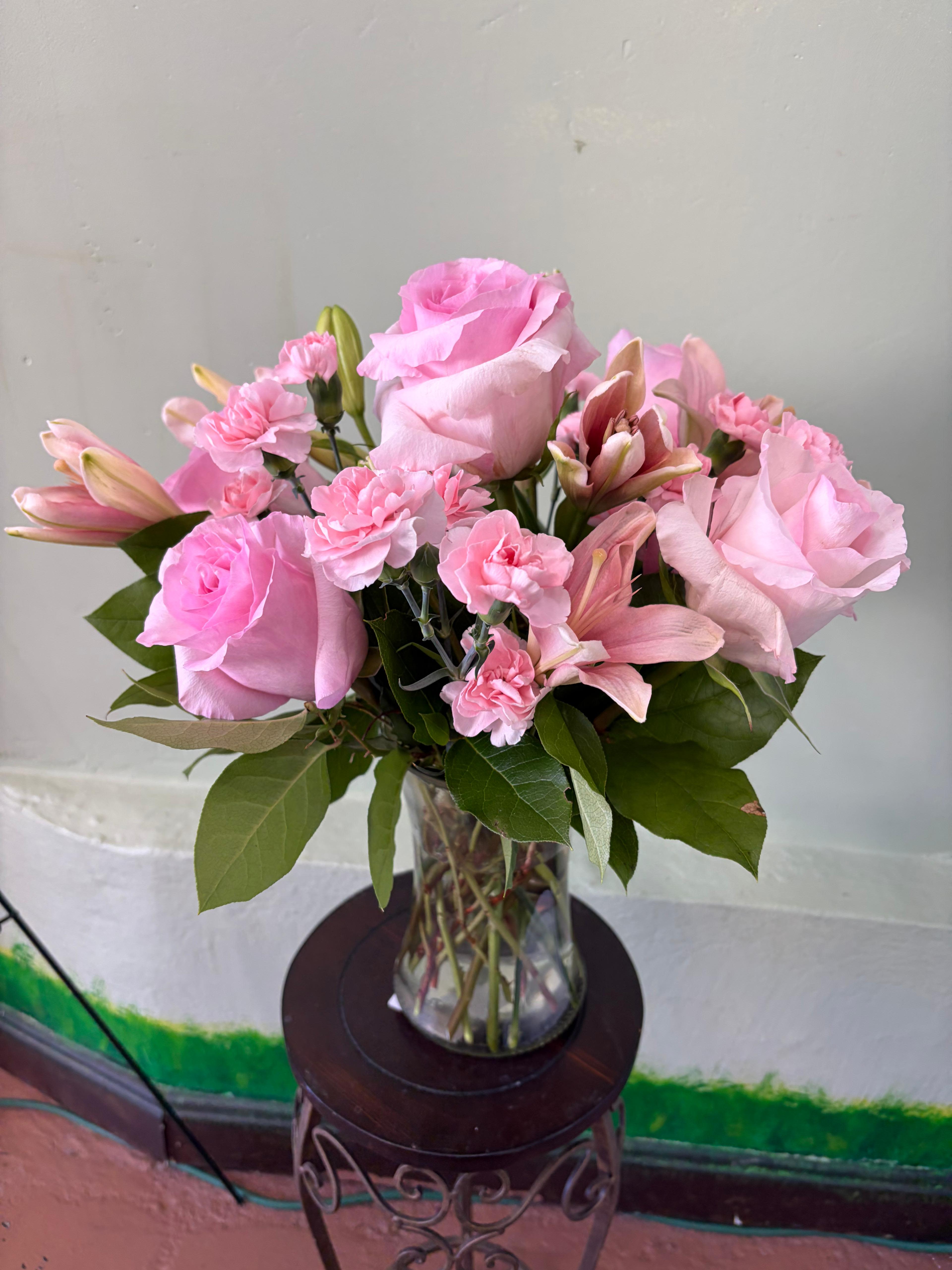 Blushing Garden Medley - Experience the timeless elegance of our Blushing Garden Medley. This soft and romantic arrangement showcases a monochromatic palette of delicate pinks, blending classic blooms with whimsical textures. Designed to evoke the feeling of a fresh-picked garden bouquet, it features velvety roses and ruffled carnations nestled among lush greenery. Housed in a classic glass gathering vase, this bouquet is the perfect gesture for anniversaries, birthdays, or simply to brighten a quiet corner of your home.